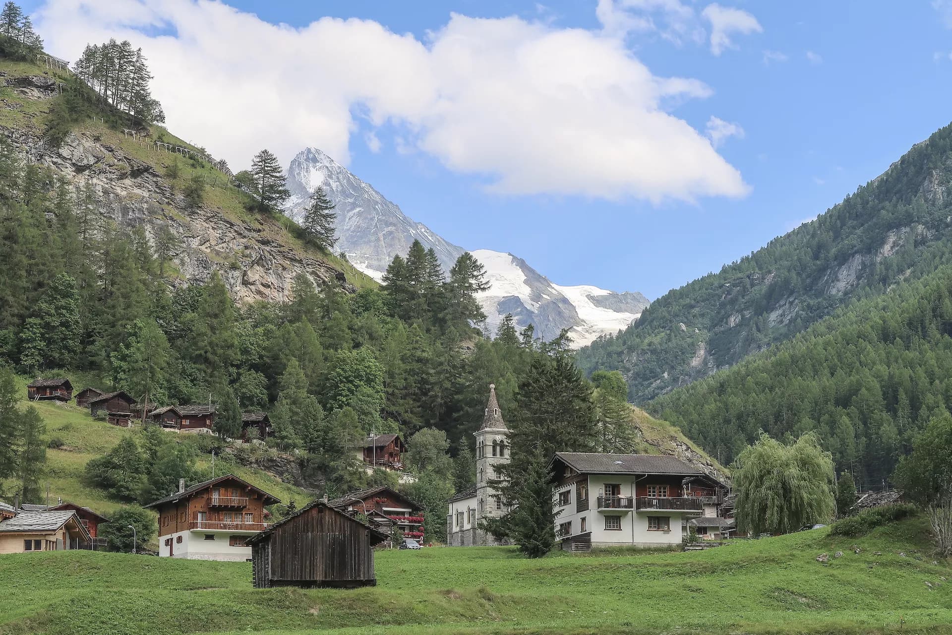 Small alpine village Les Hauderes nestled in green valley with snow-capped mountains under blue sky.