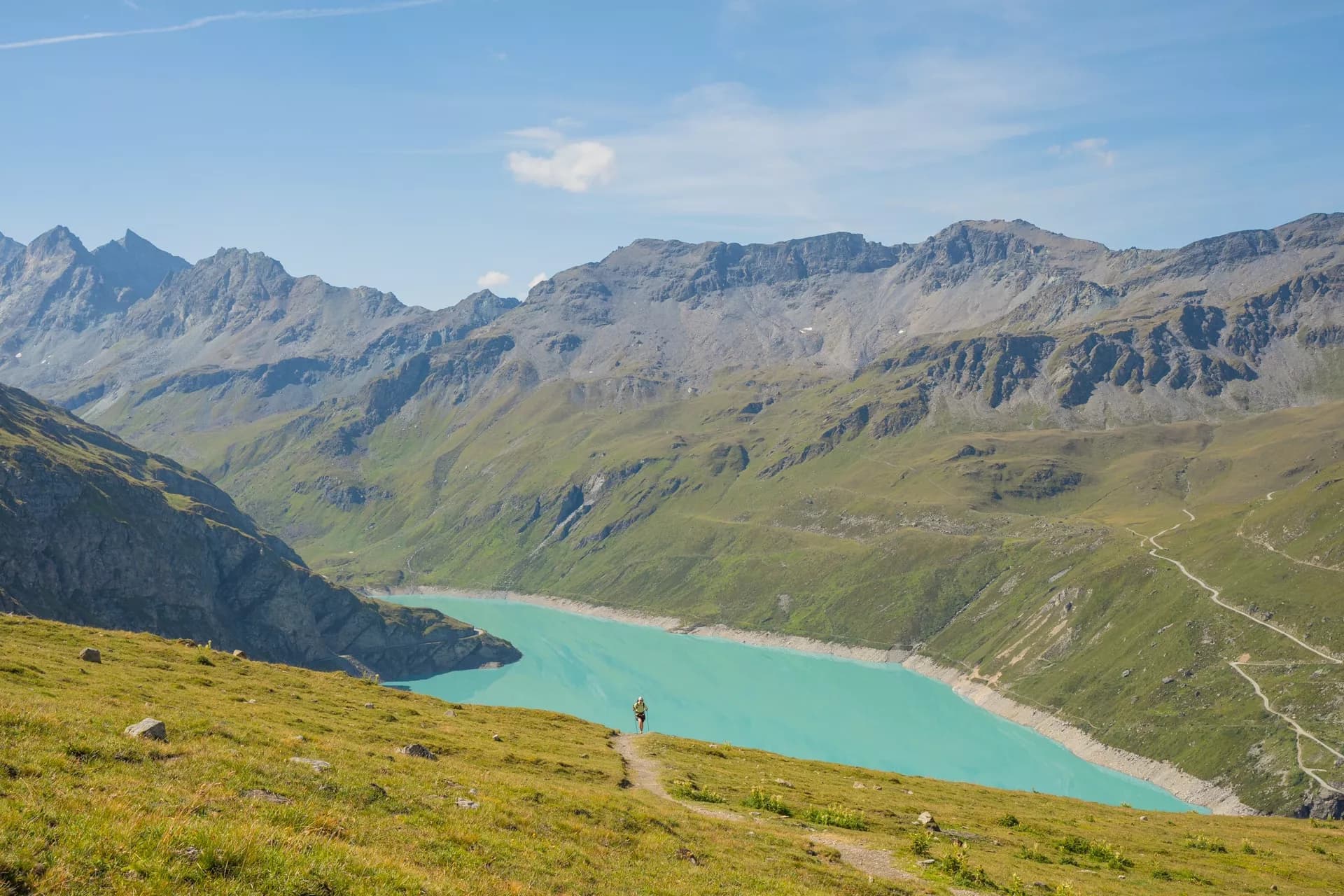 Hiker on grassy trail overlooking turquoise reservoir surrounded by rugged green and gray mountains.
