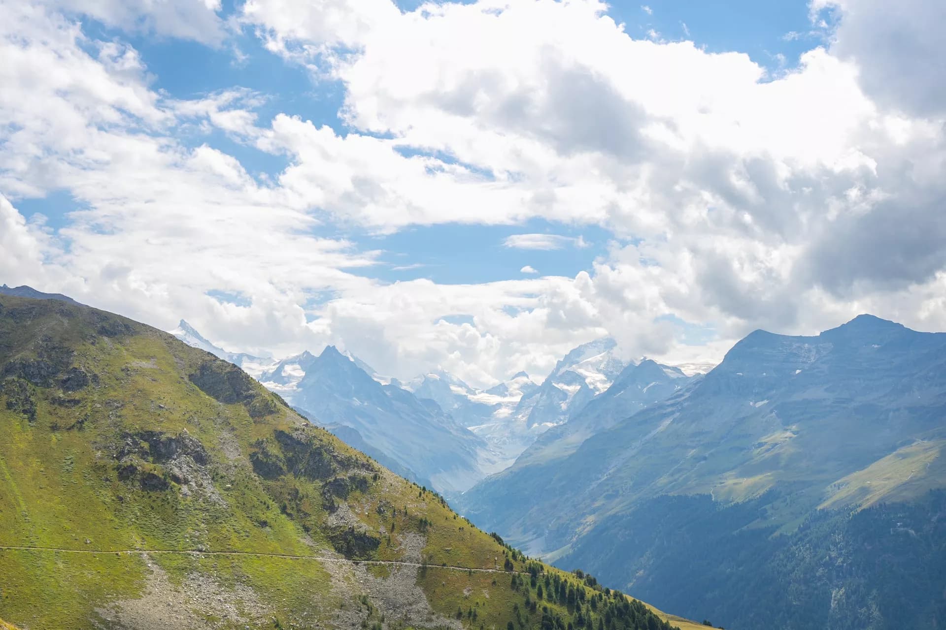 Mountain valley view with green slopes, distant snow-capped peaks, and dramatic clouds.