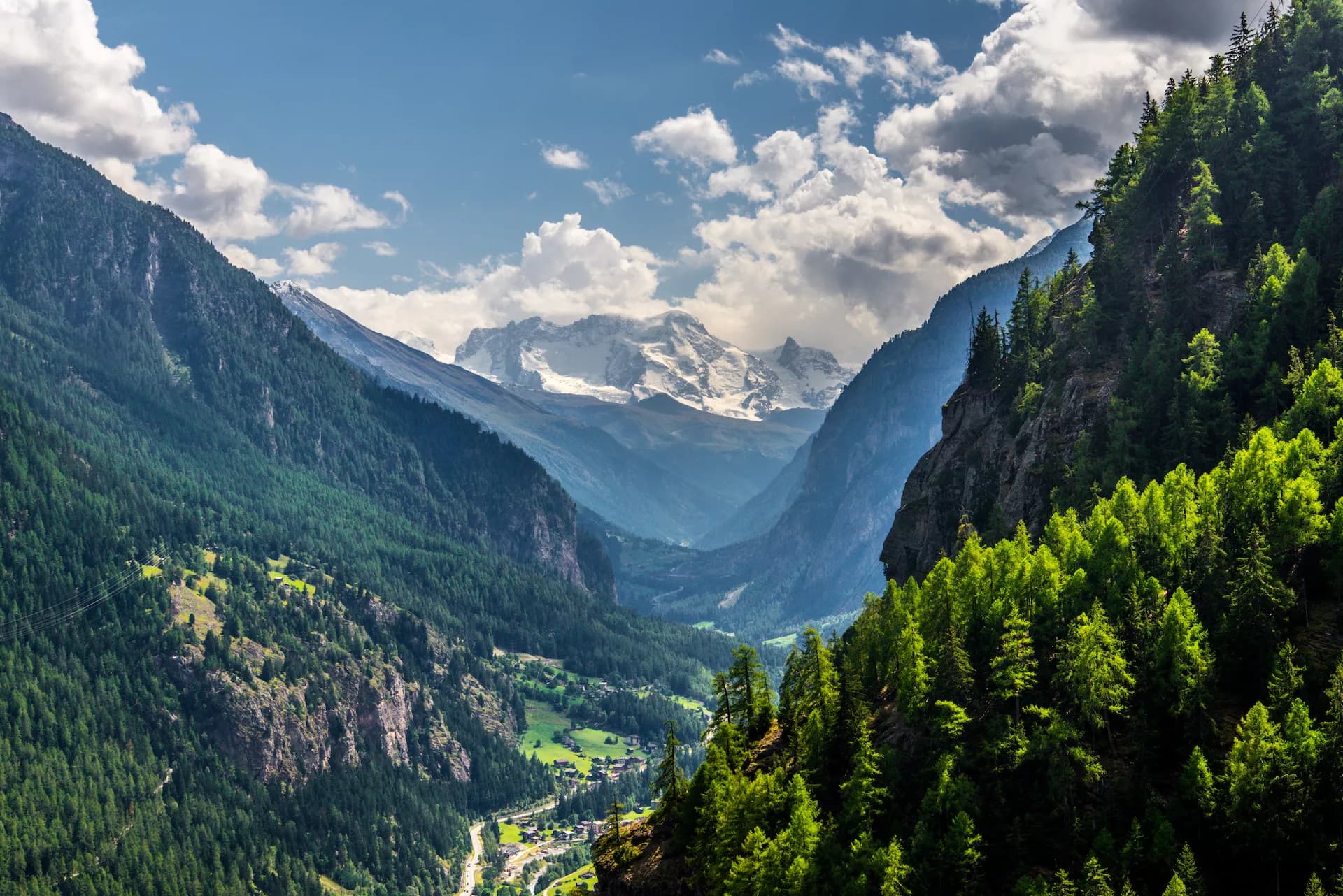 Alpine valley in Mattertal, Switzerland with steep forested slopes and snow-capped peaks under a cloudy sky.