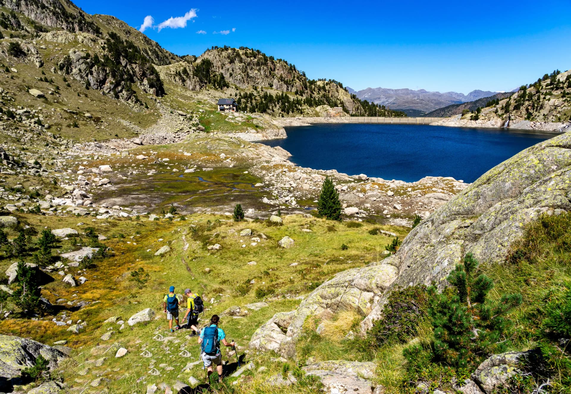 Hikers ascending grassy slope toward a bright blue alpine lake with mountains and a dam.