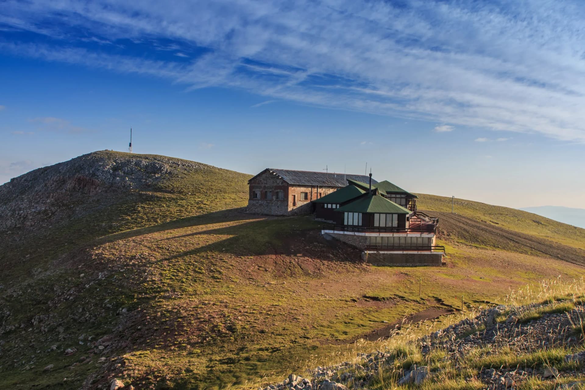 Mountain refuge building with green roof on grassy slope under blue sky with wispy clouds