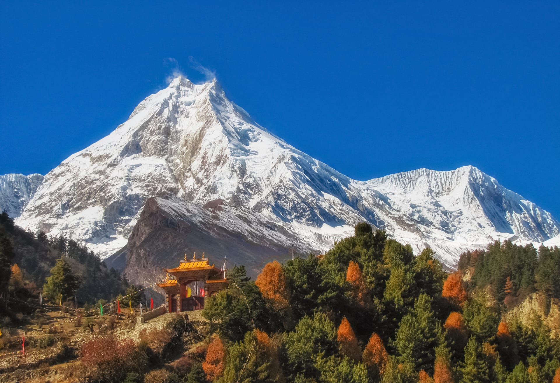Buddhist monastery gate below massive snow-covered Manaslu mountain peak under clear blue sky.