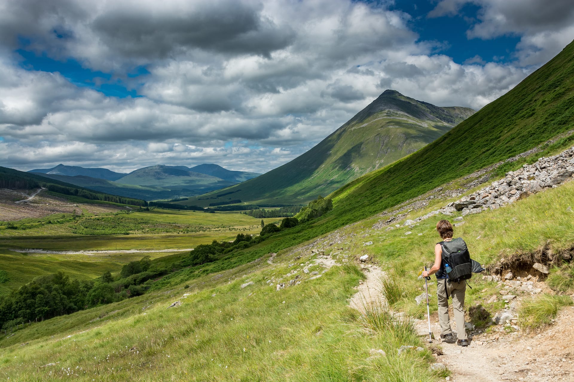 Hiker with backpack and trekking pole ascending grassy mountain trail under cloudy sky.