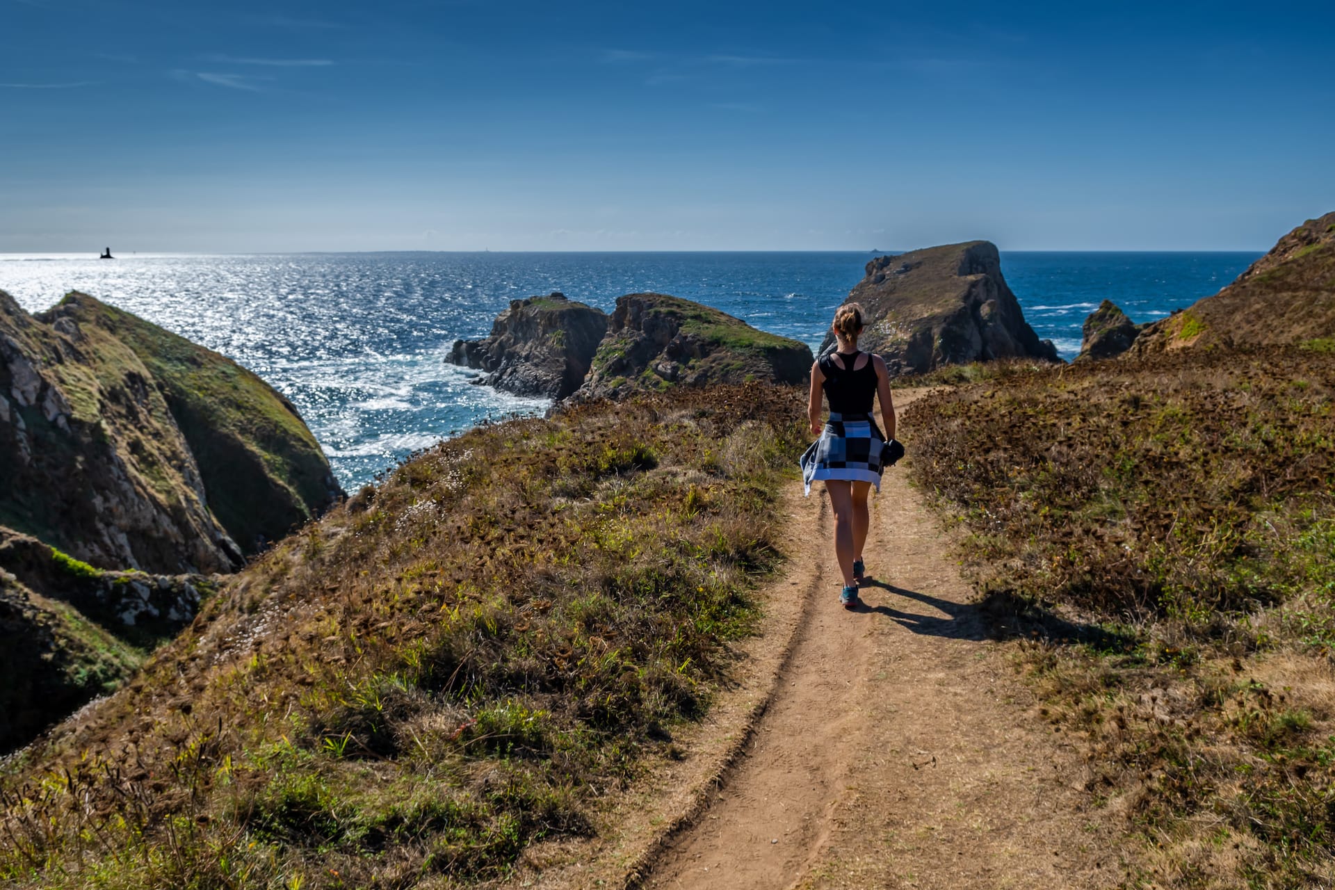 Woman hiking on dirt path along rugged coastal cliffs overlooking sparkling blue ocean water.