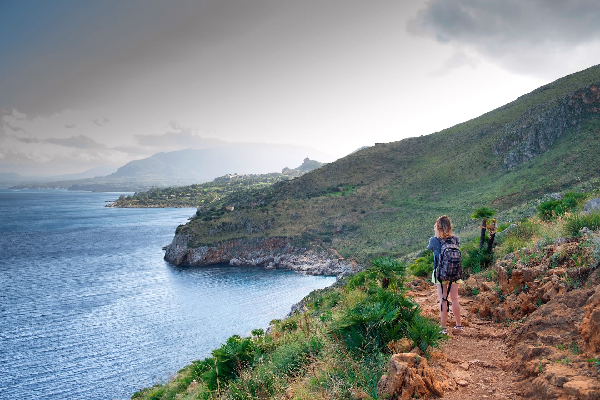 Hiker with backpack on rocky coastal trail overlooking blue Mediterranean sea and green hills.