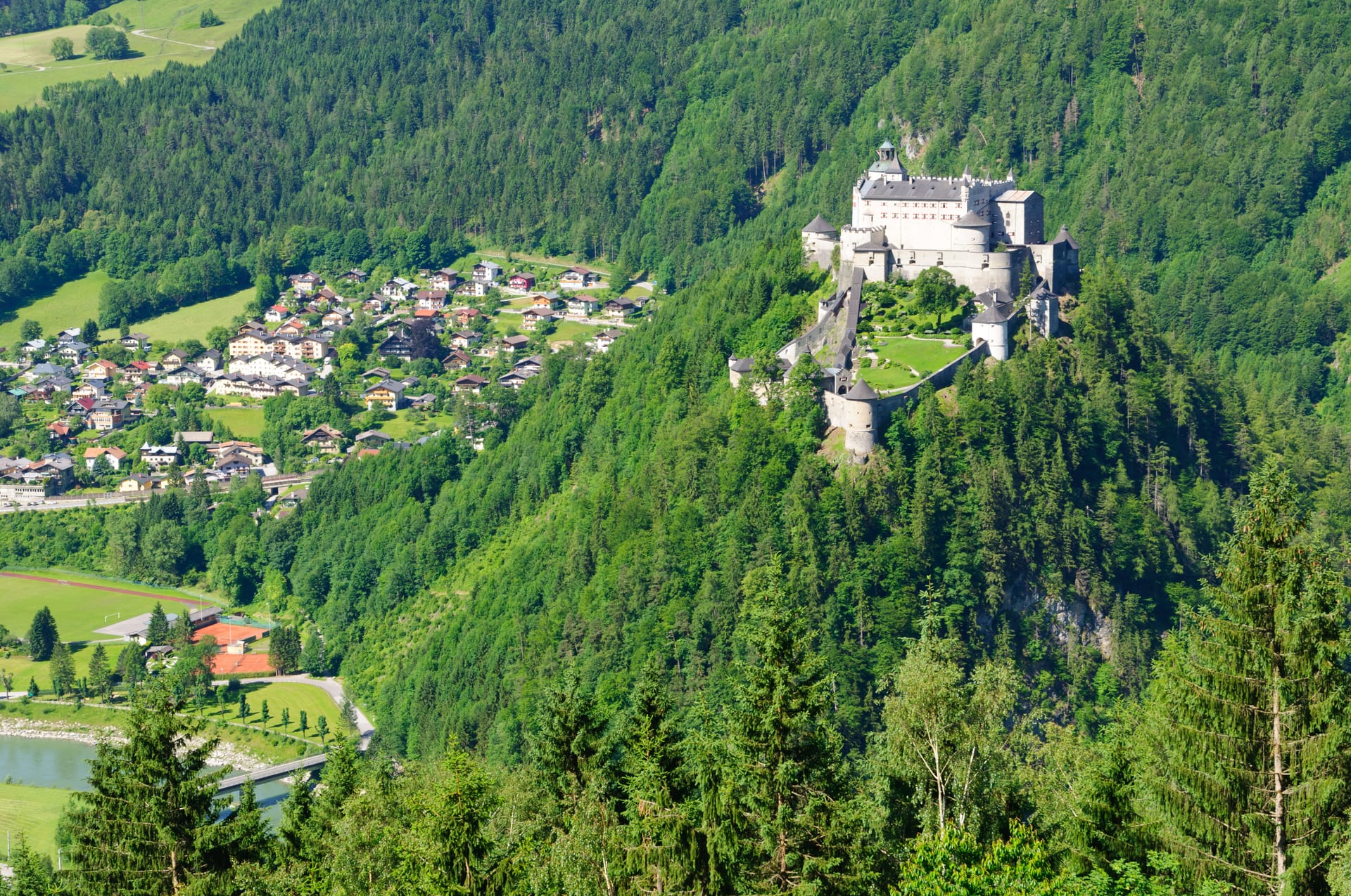 Castle on forested mountain overlooking alpine village and river valley in summer.