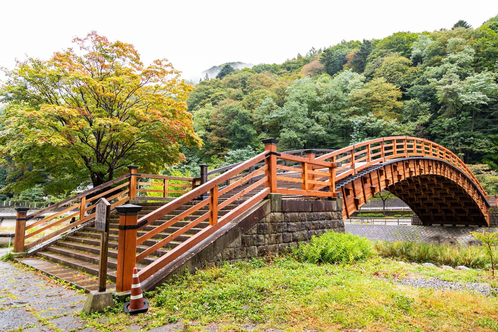 Arched wooden bridge with stairs leading up, set against a lush, forested hillside in Japan.