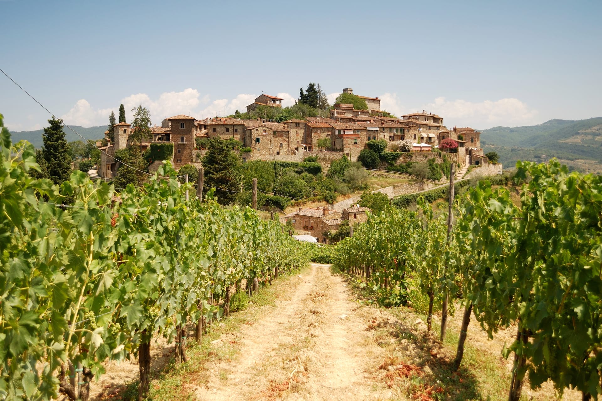 Vineyard path leading to historic stone village on hillside under blue sky in Tuscany.