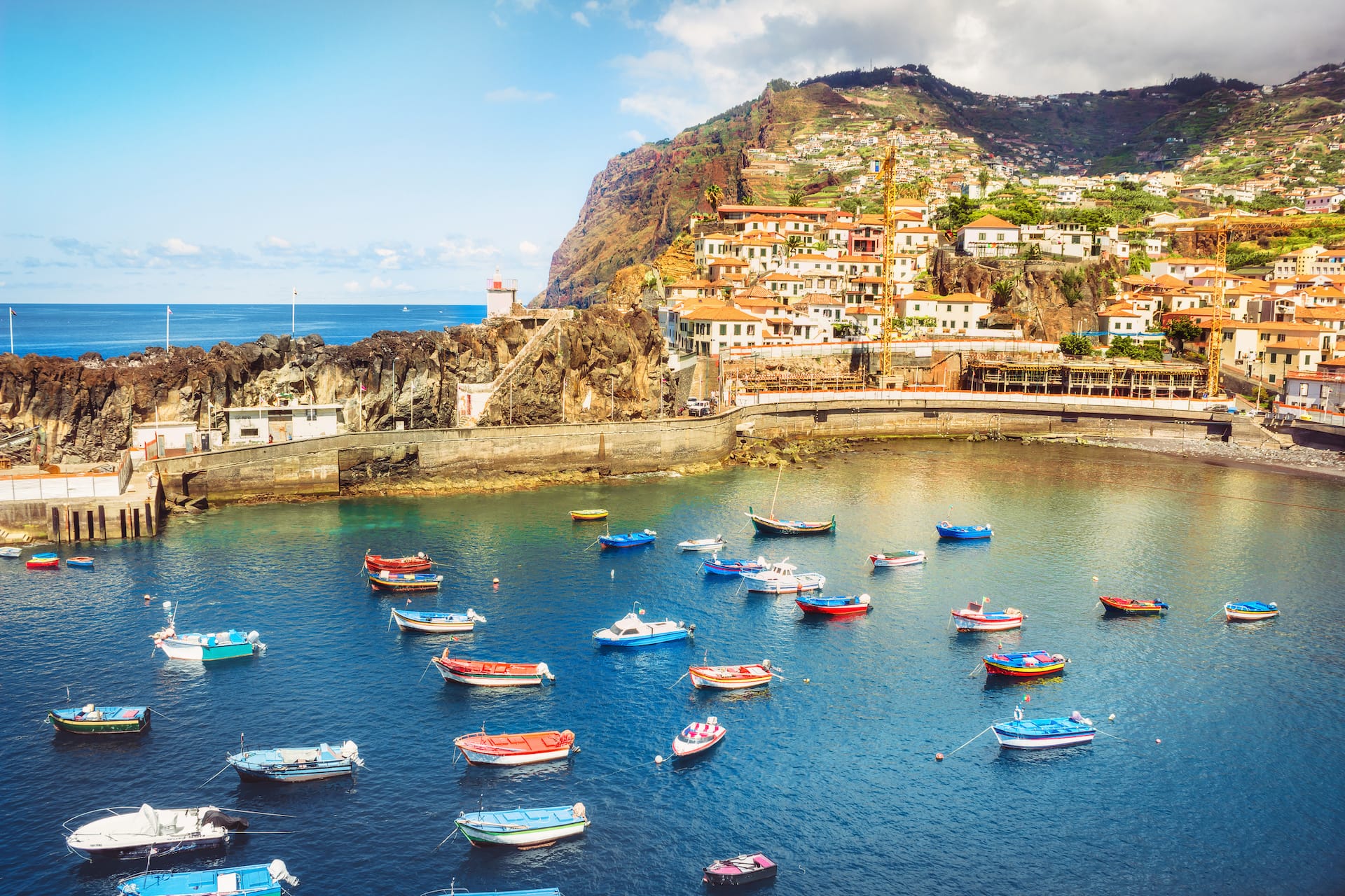 Colorful fishing boats moored in a harbor below a coastal town built into a steep hillside.