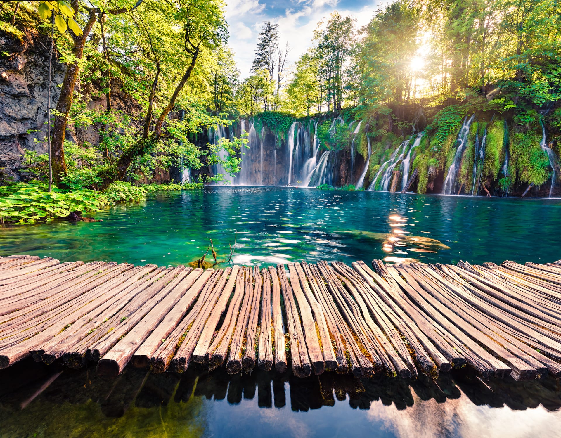 Wooden boardwalk over turquoise lake beneath waterfalls and lush green forest in Croatia.