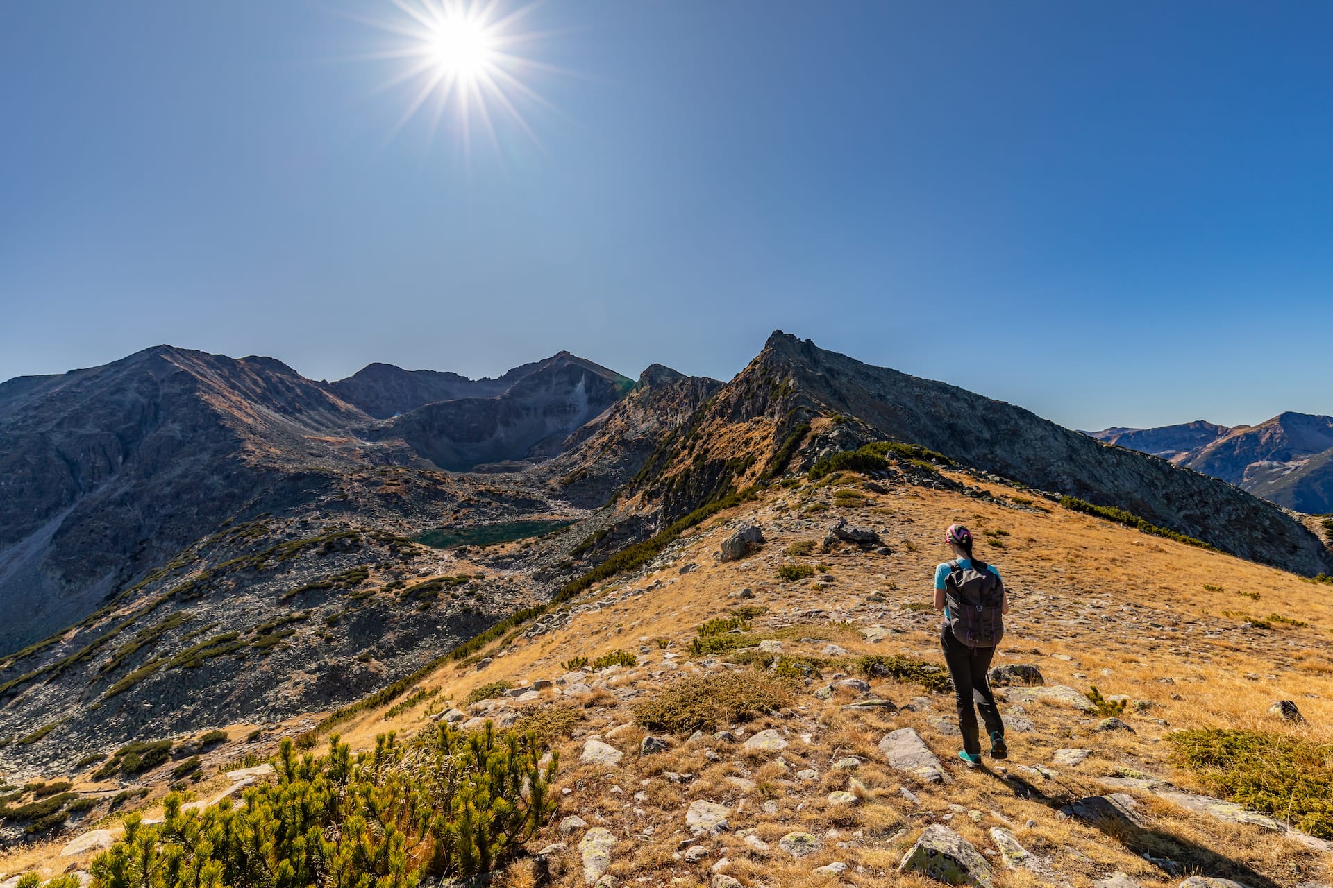 Hiker walking on grassy ridge overlooking rugged mountains and small alpine lake under bright sun.