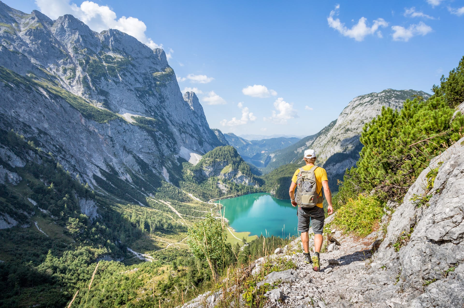 Hiker with backpack walking on rocky trail overlooking turquoise alpine lake and steep mountains