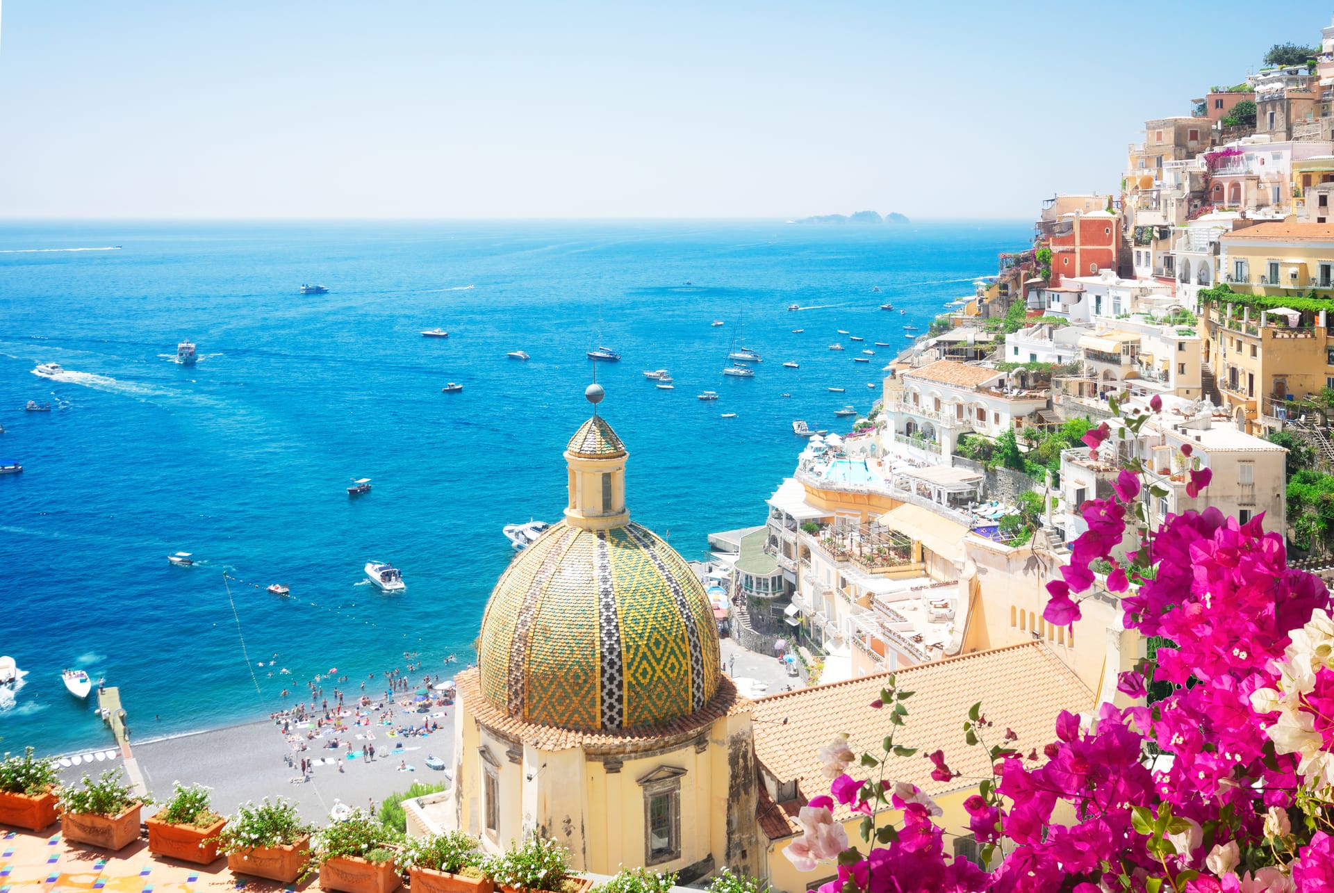 Colorful tiled dome church overlooking Amalfi Coast town, beach, and blue sea with boats.