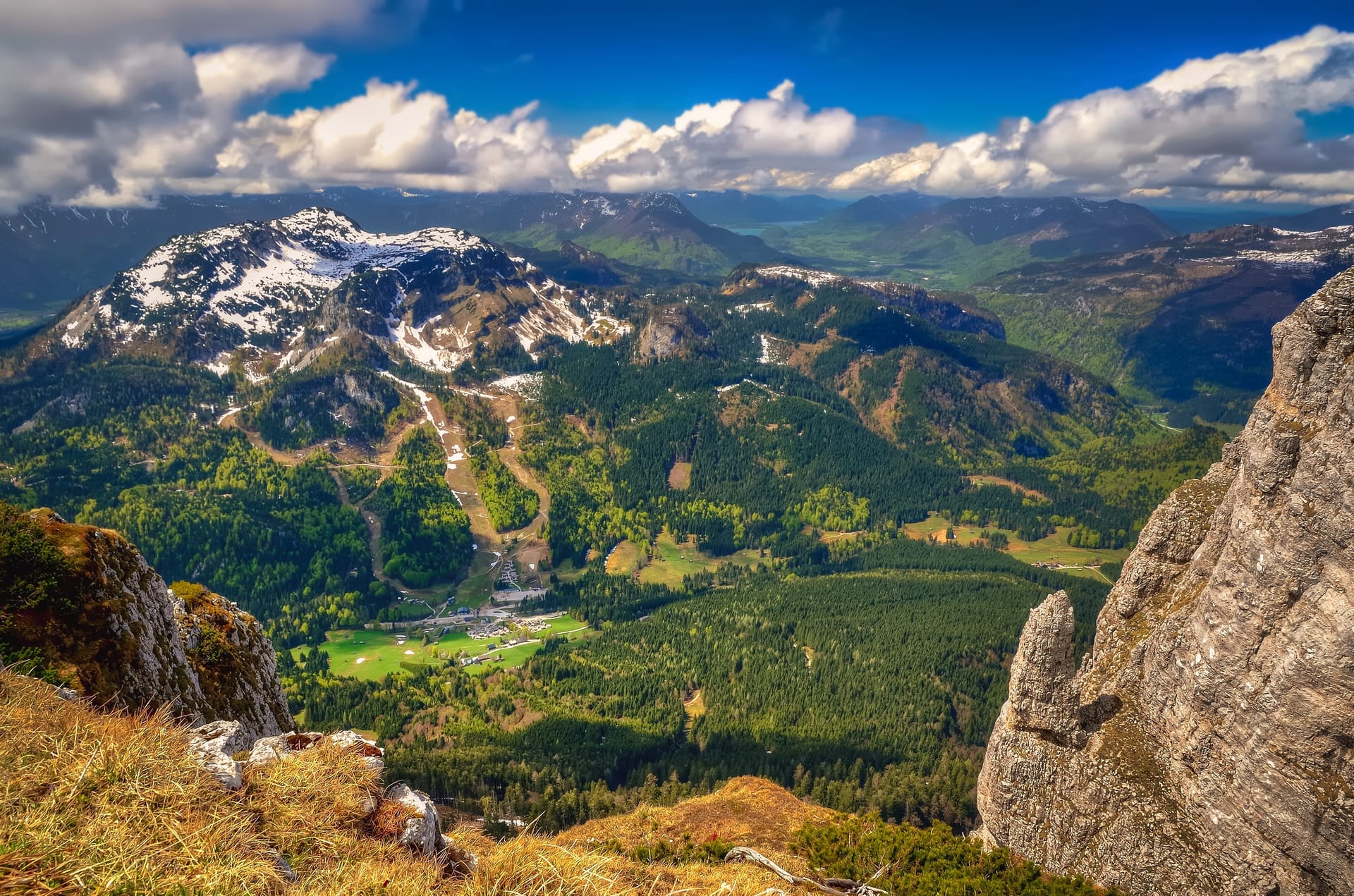 Mountain vista from rocky overlook with snow-capped peaks, green valleys, and blue sky with clouds.