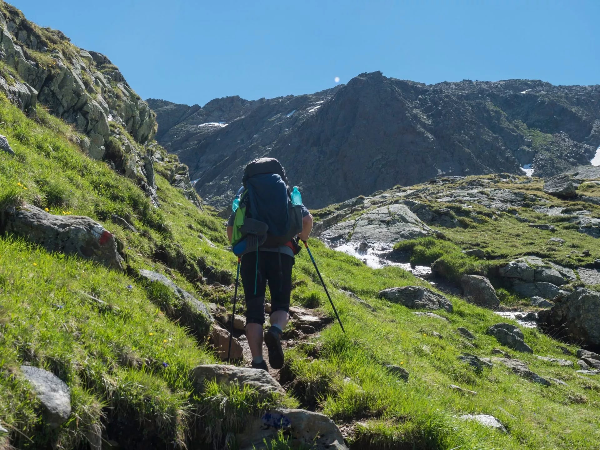 Lonely man hiker with heavy backpack at Stubai hiking trail, Stubai Hohenweg at green summer alpine mountain valley with spring stream. Tyrol, Austrian Alps