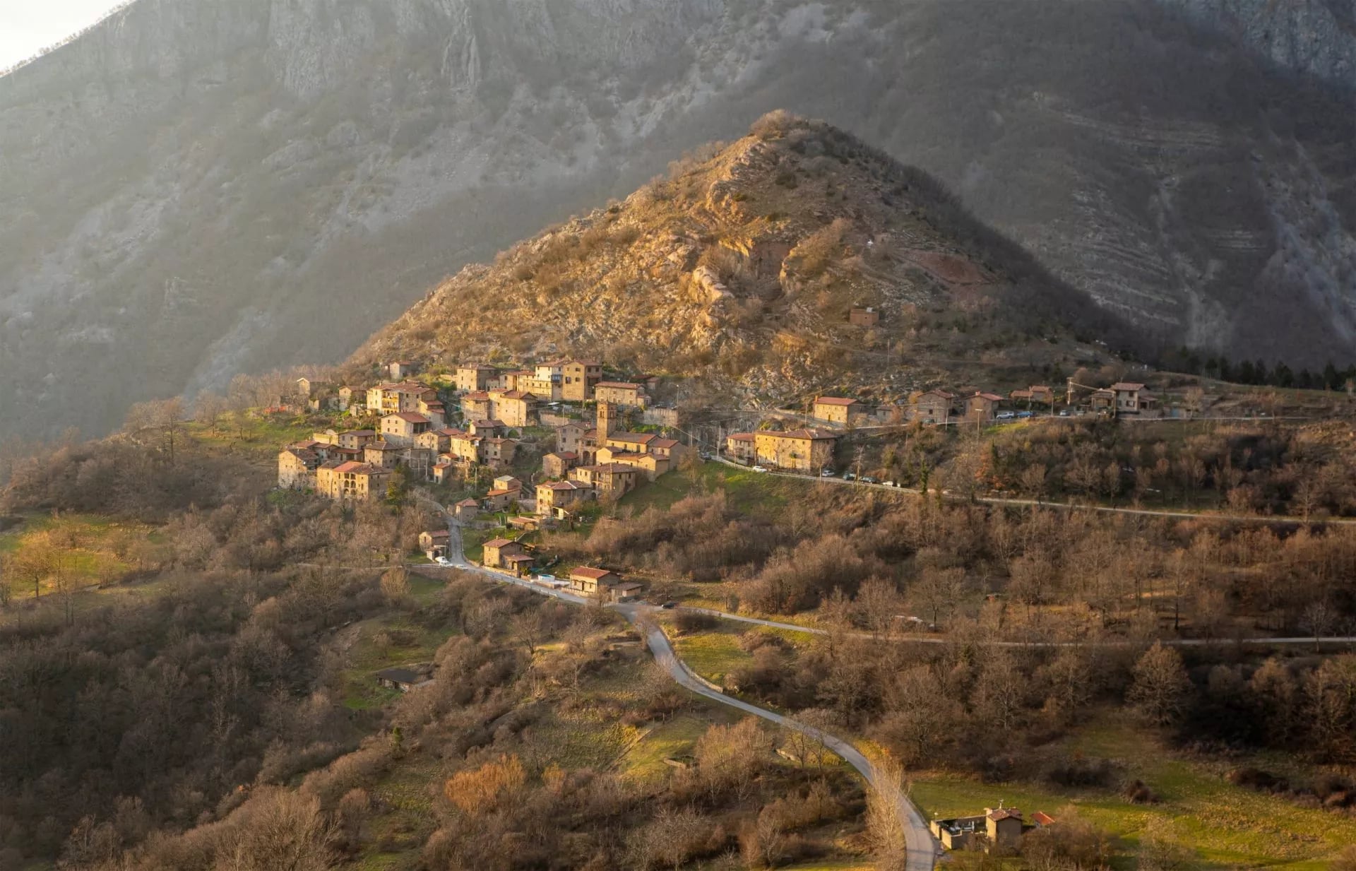 Hillside village of stone houses nestled on a sunlit slope below large mountains, Pania di Corfino.