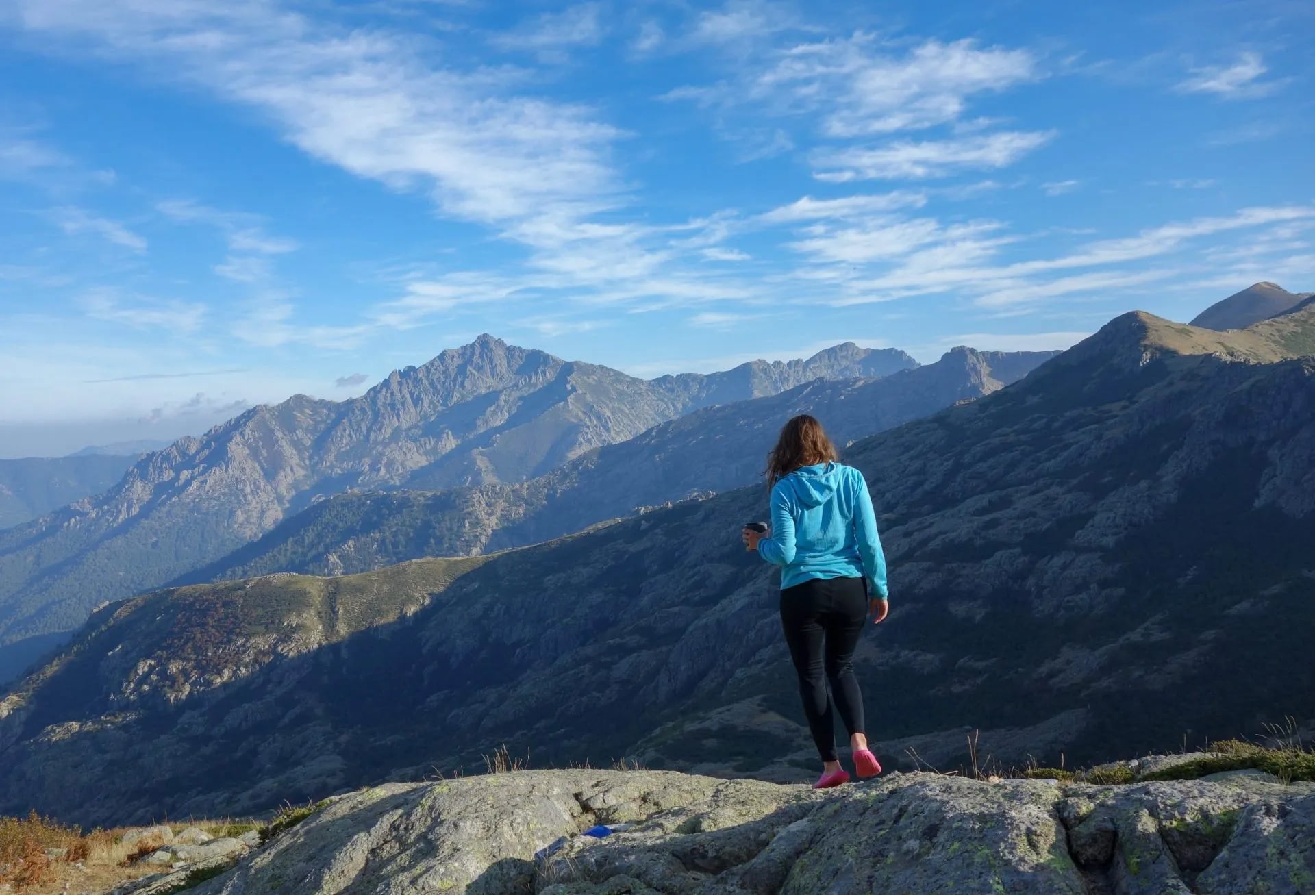 Hiker standing on rocks overlooking rugged mountains under a blue sky in Corsica.