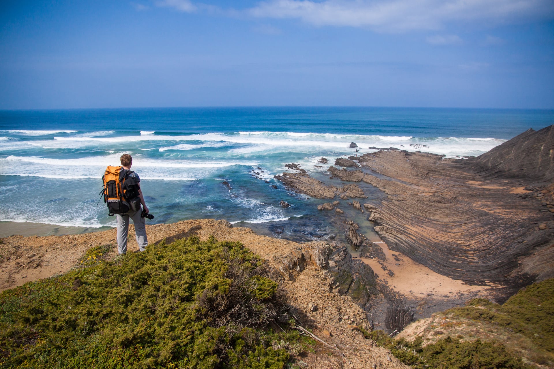 Hiker with backpack overlooking Rota Vicentina coastline with layered rock formations and ocean waves.