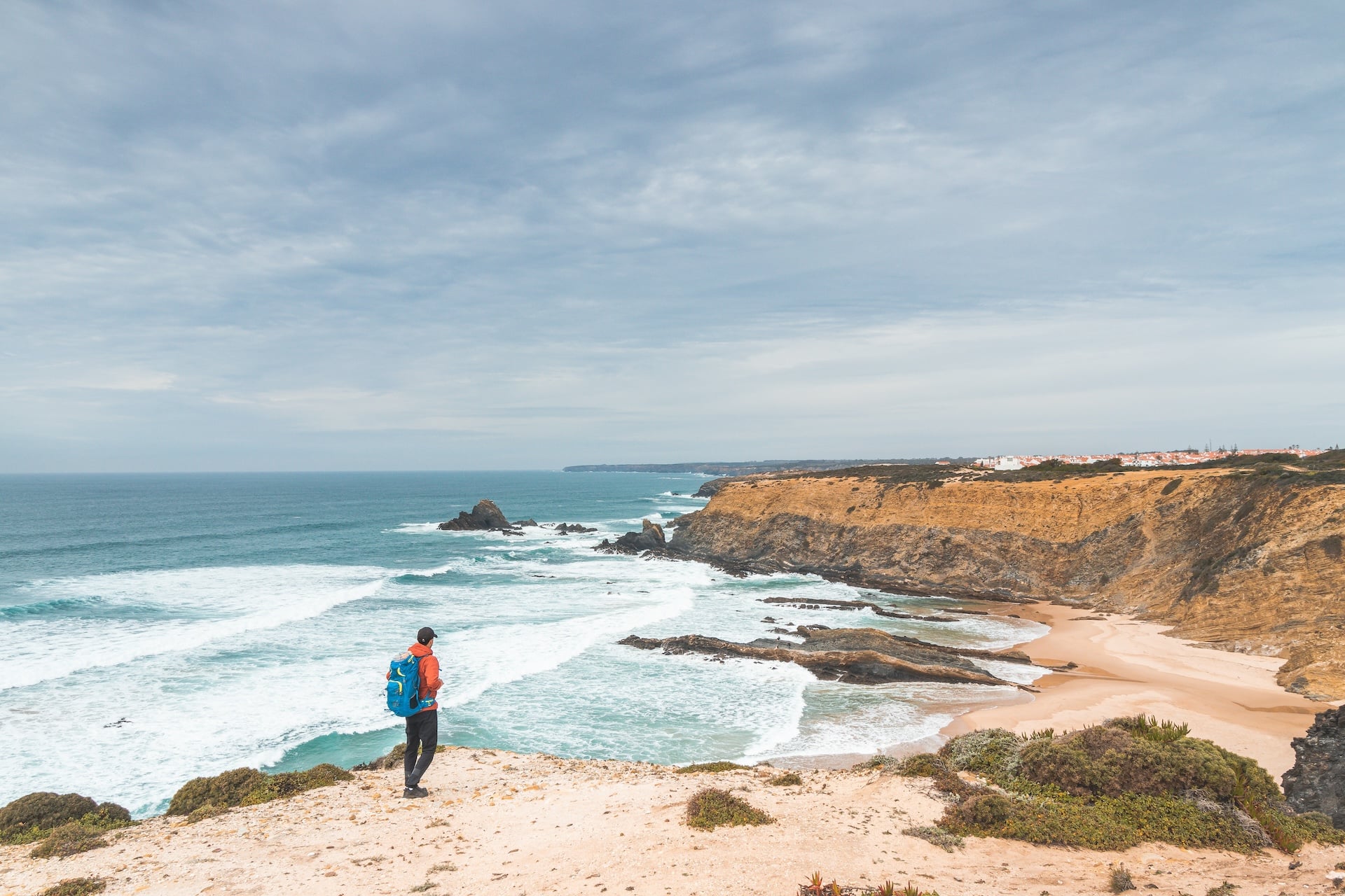 Hiker with blue backpack overlooking rugged coastline, waves, and beach in Zambujeira do Mar.