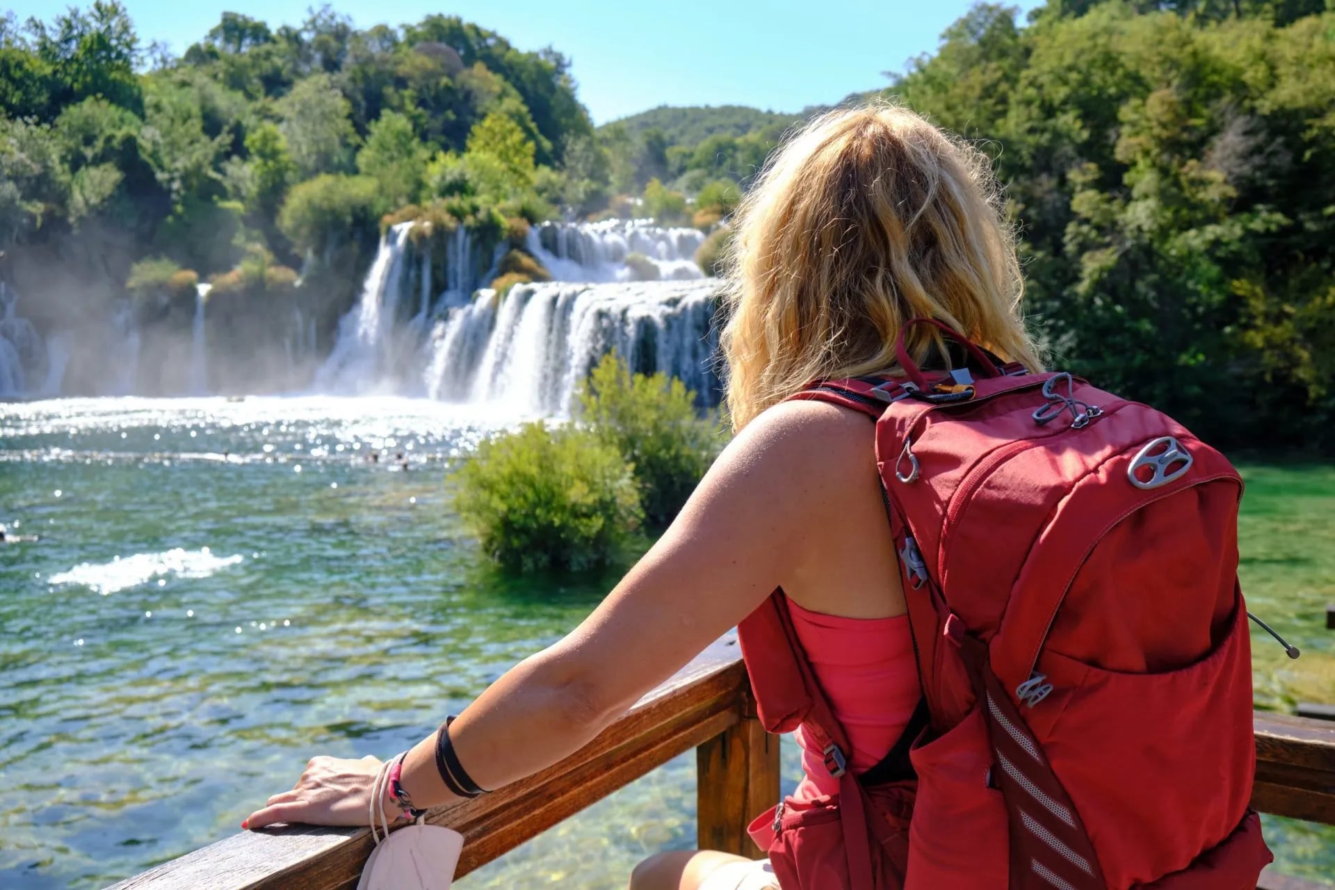 Hiker with red backpack viewing waterfalls cascading into sparkling green water at Krka National Park.