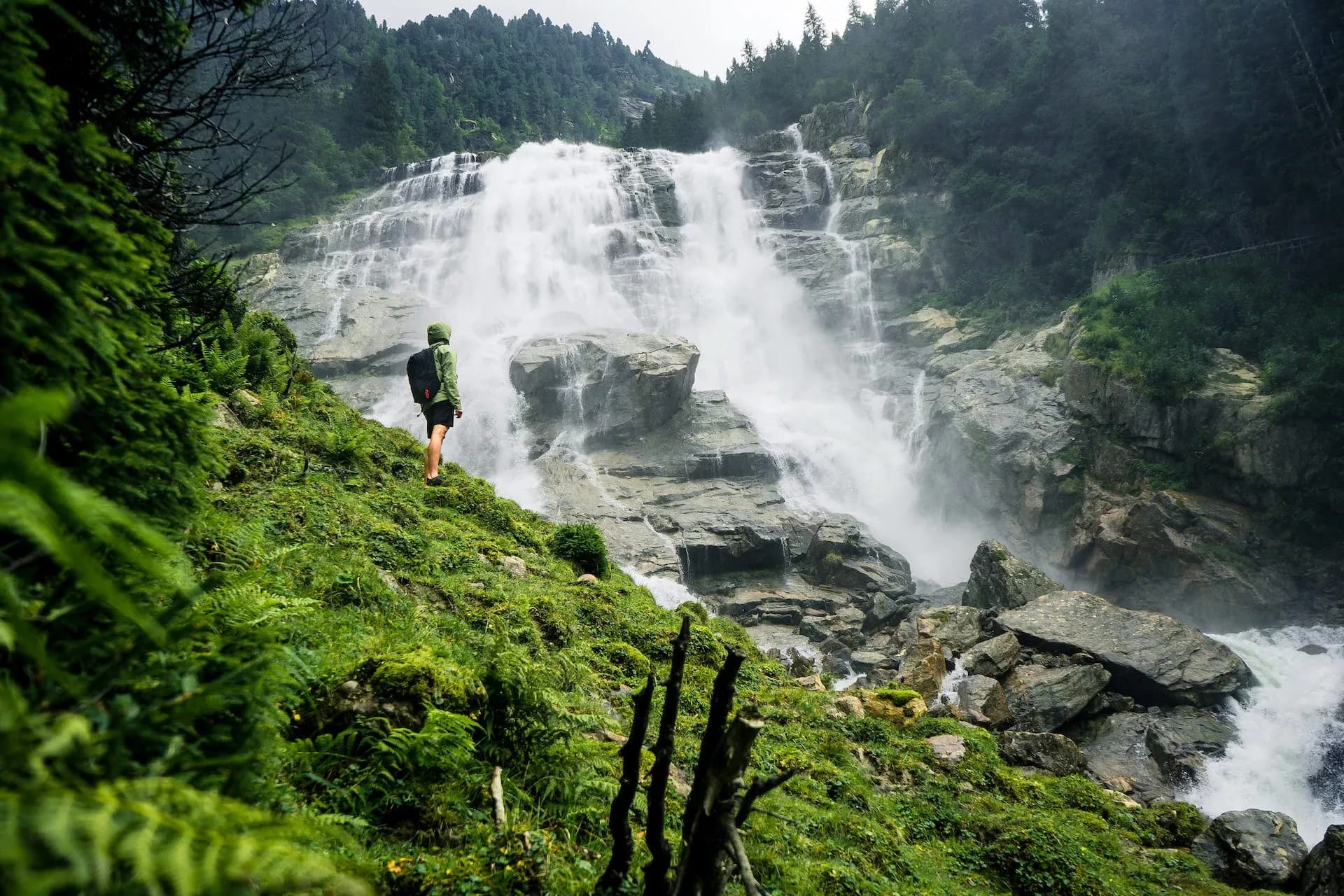 Hiker observing the powerful Grawa Waterfall cascading down mossy rocks surrounded by dense green forest.