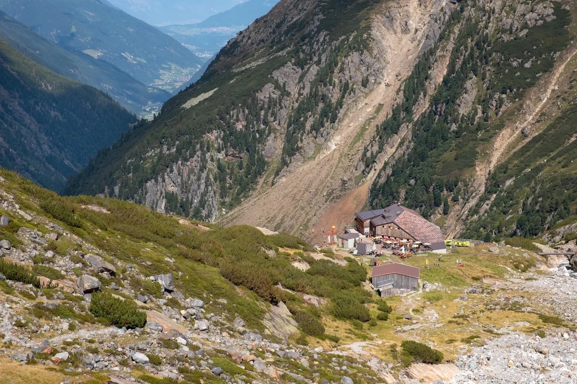 Alpine mountain hut with outdoor seating nestled in a steep valley with distant town view