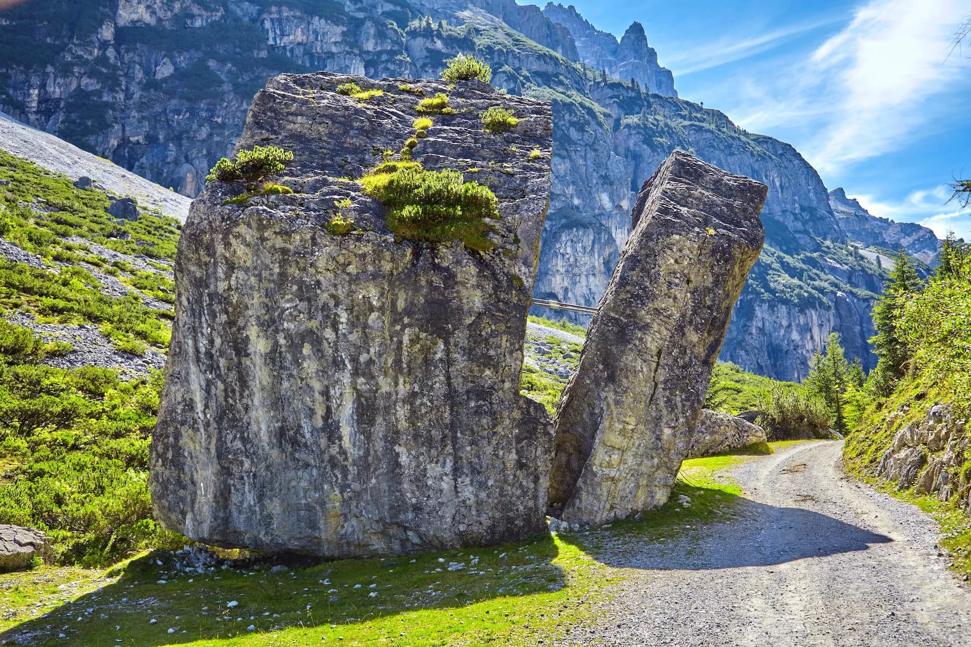 Large split boulder next to a gravel path with steep, green mountains under a blue sky.