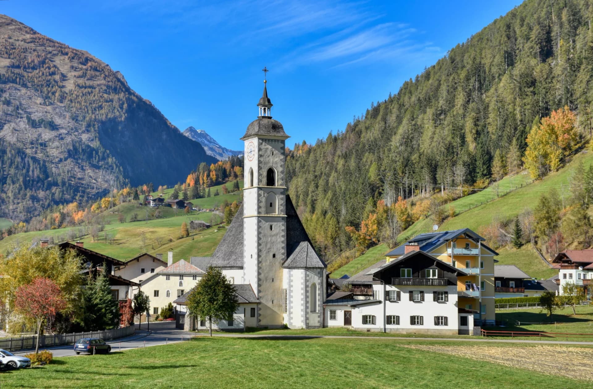 Alpine village church with bell tower set against steep, forested mountainsides in autumn.