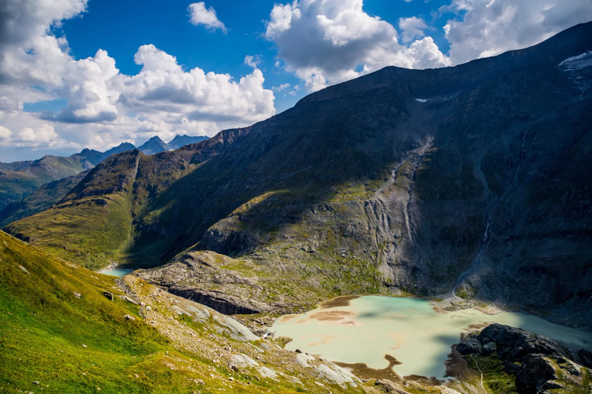 Glacial lake below Mount Grossglockner with steep green and rocky slopes under a blue sky.