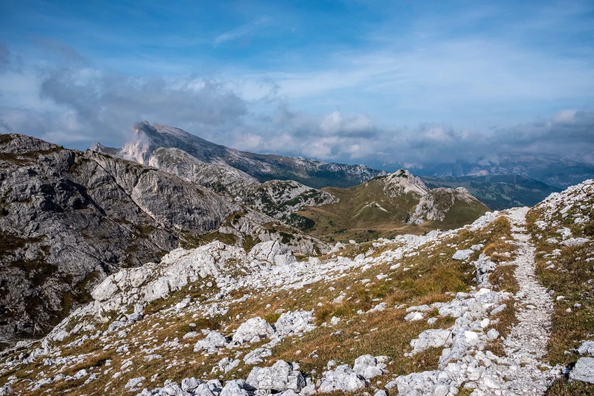 Hiking trail over rocky alpine terrain with distant mountains under a cloudy blue sky, Valparola.