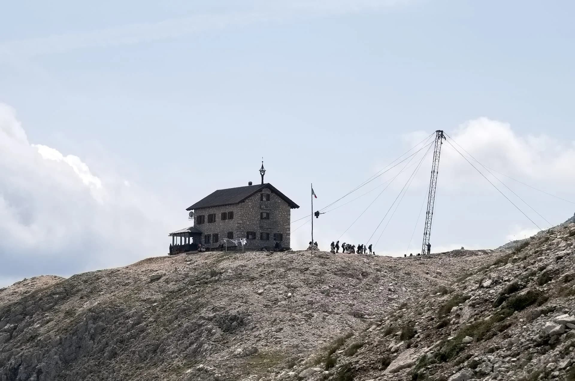 Stone mountain hut with hikers and radio tower on rocky summit under cloudy sky