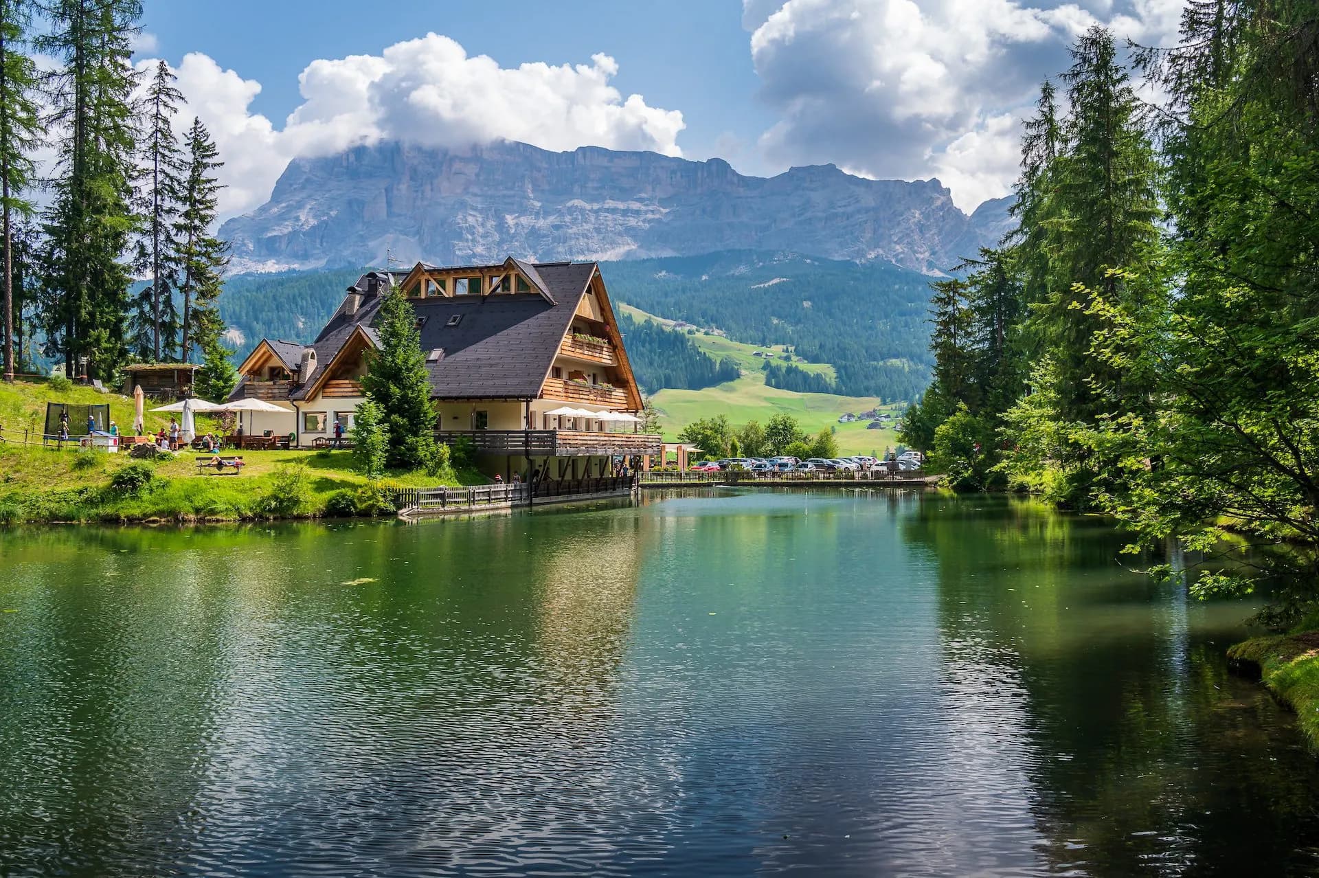 Alpine lake with wooden lodge, green hills, and towering mountains under a cloudy sky.