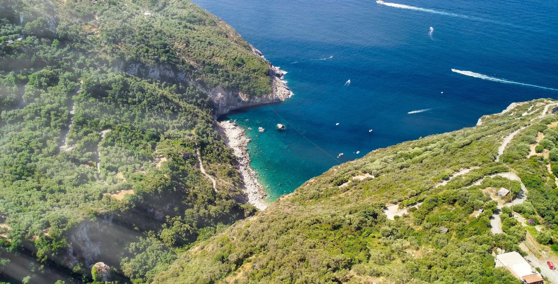 Aerial view of heavily wooded cliffs overlooking a small cove with turquoise water and boats near Sorrento.