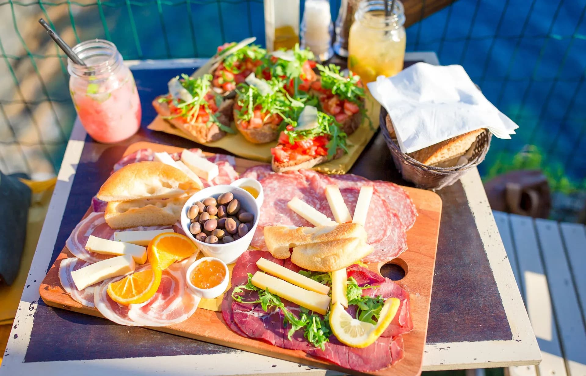Charcuterie board with meats, cheese, olives, bruschetta, and drinks outdoors by blue water.