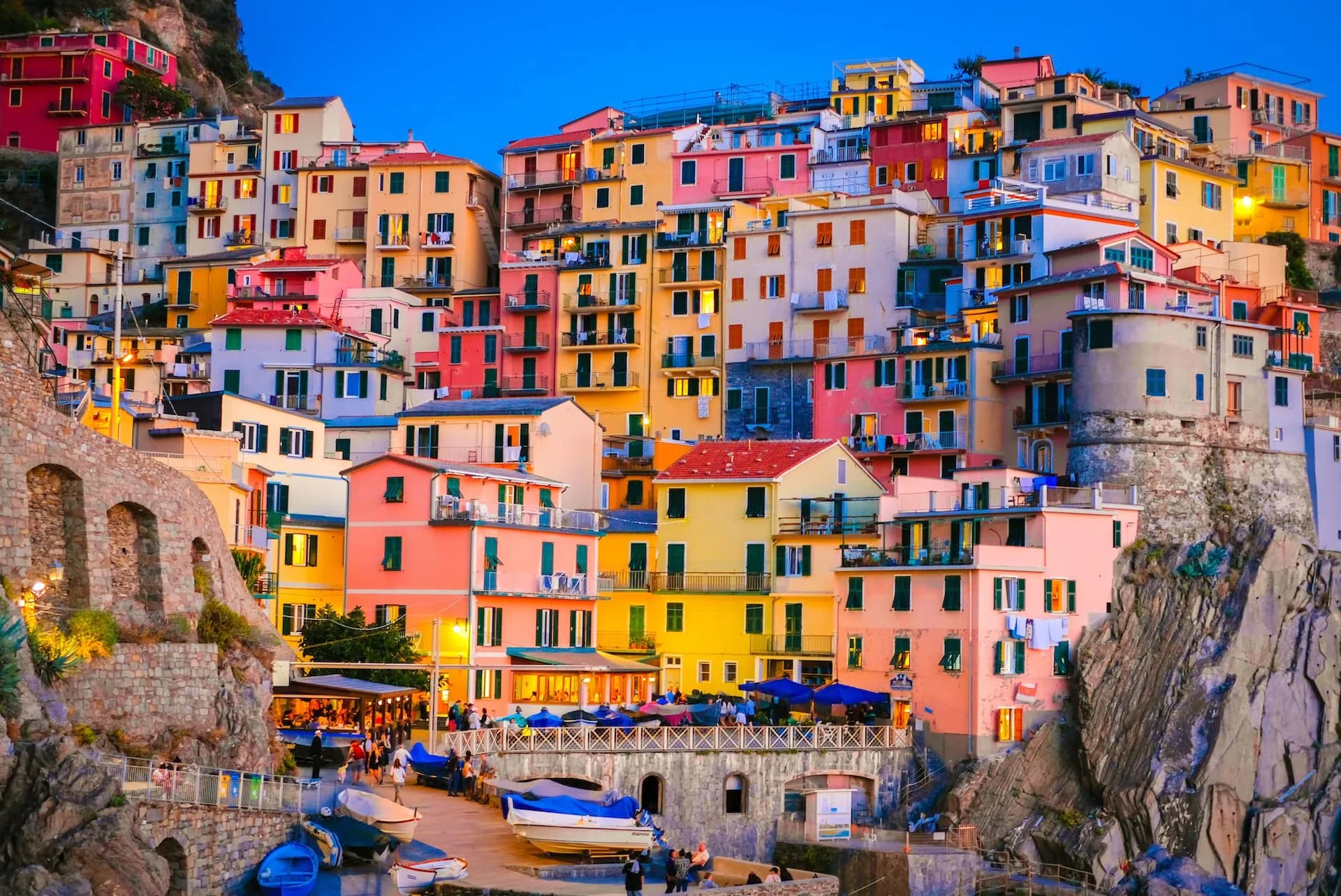 Colorful stacked houses clinging to a cliffside above a harbor with boats at dusk in Manarola.