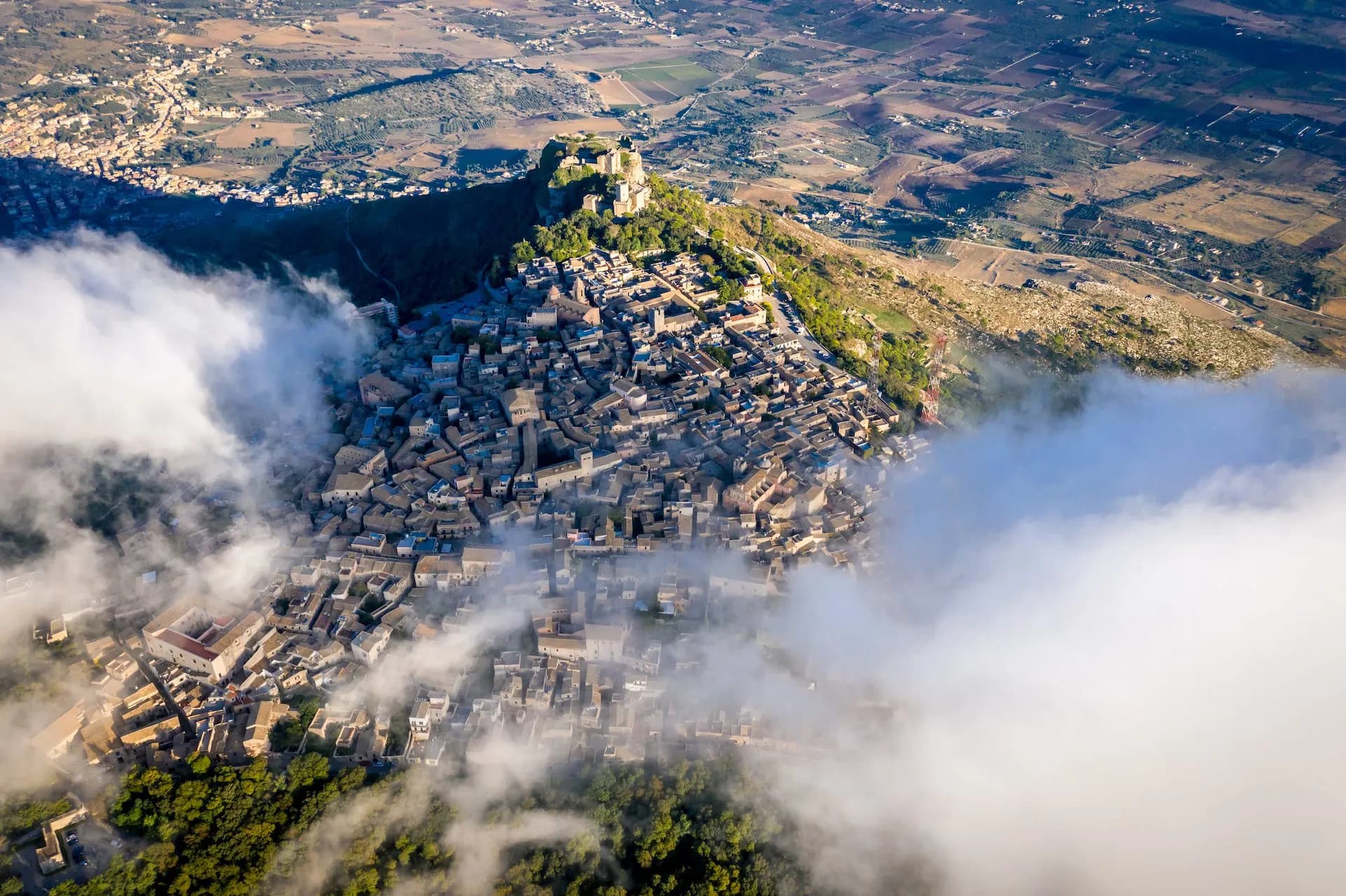 Aerial view of Erice town nestled on a hilltop with a castle, partially obscured by clouds.