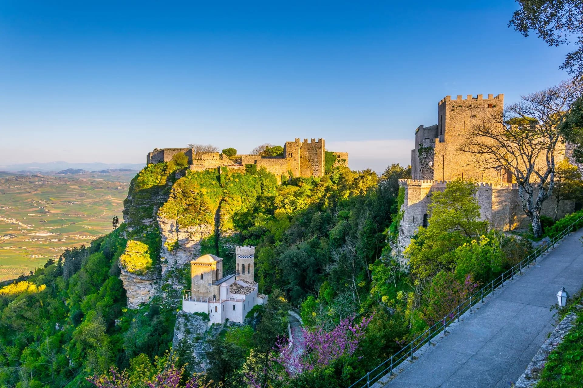 Erice Castle ruins atop a lush cliff overlooking a green valley under a clear blue sky.
