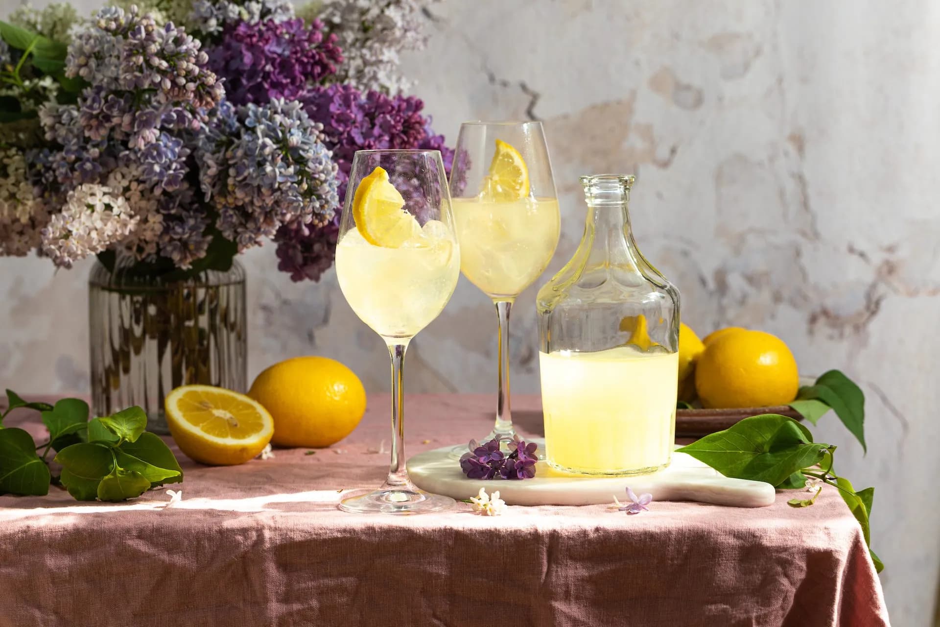 Two glasses of fresh lemonade with lemon slices, a bottle, and lemons on a pink cloth with lilacs.