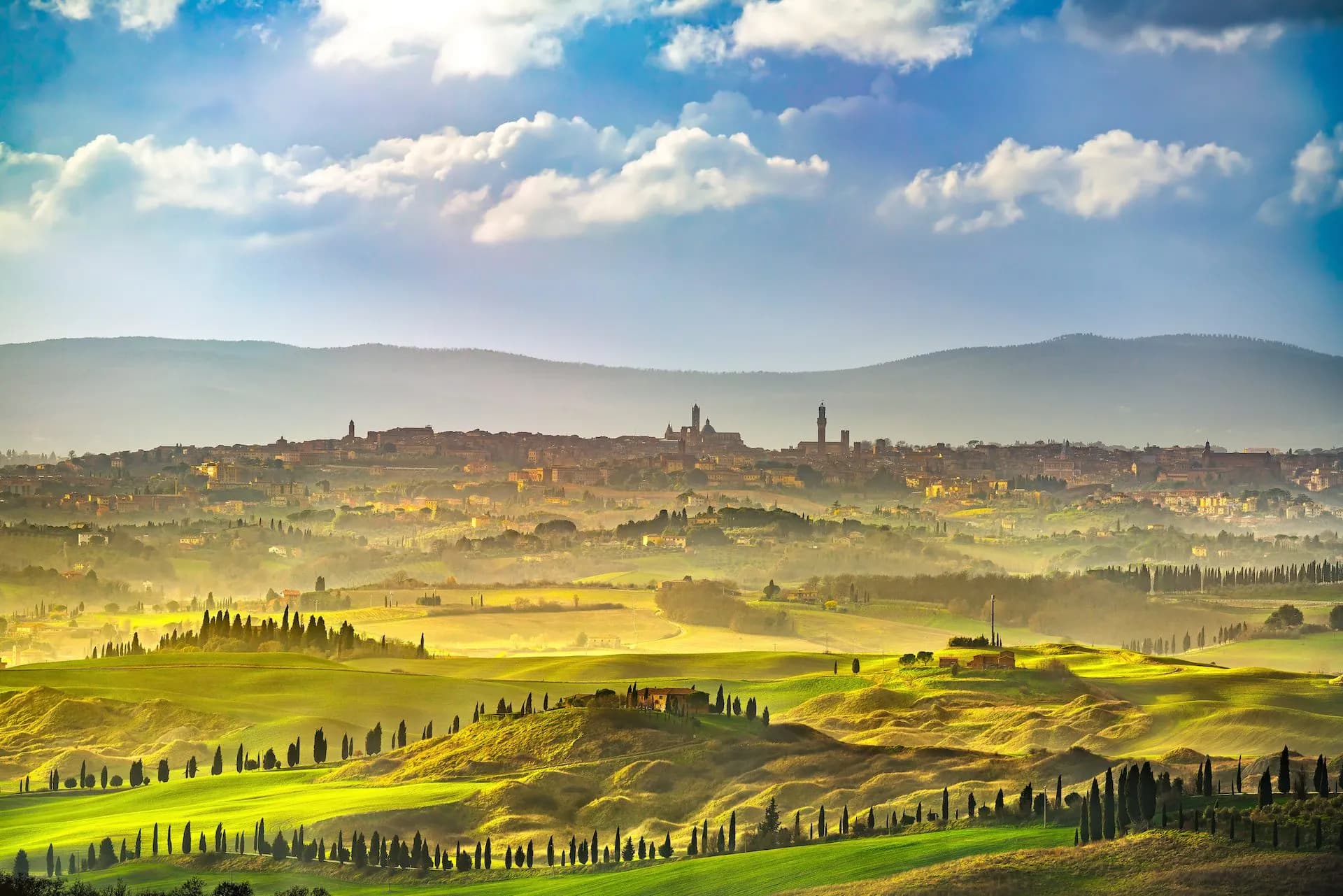 View of Siena cityscape over rolling green Tuscan hills with cypress trees under a cloudy blue sky.