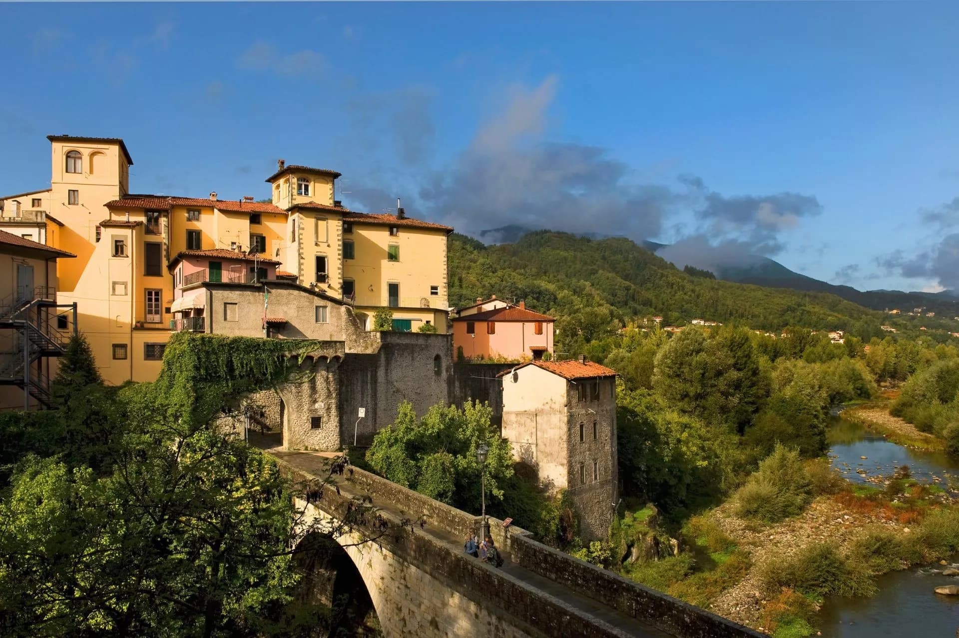 Historic buildings above stone bridge crossing river in Castelnuovo di Garfagnana with green mountains.