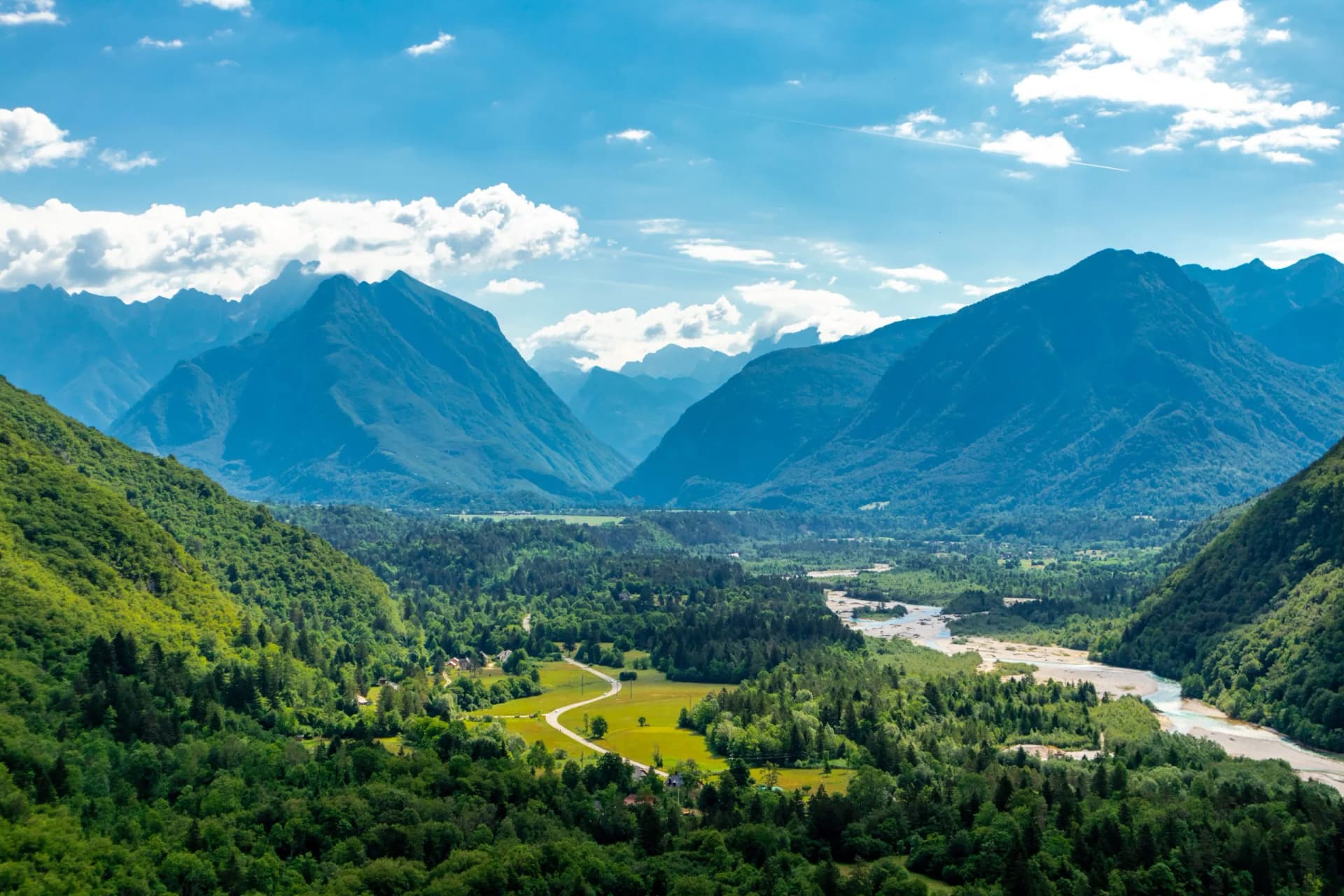 Lush green valley with winding river, dense forest, and steep blue mountains under a cloudy sky.