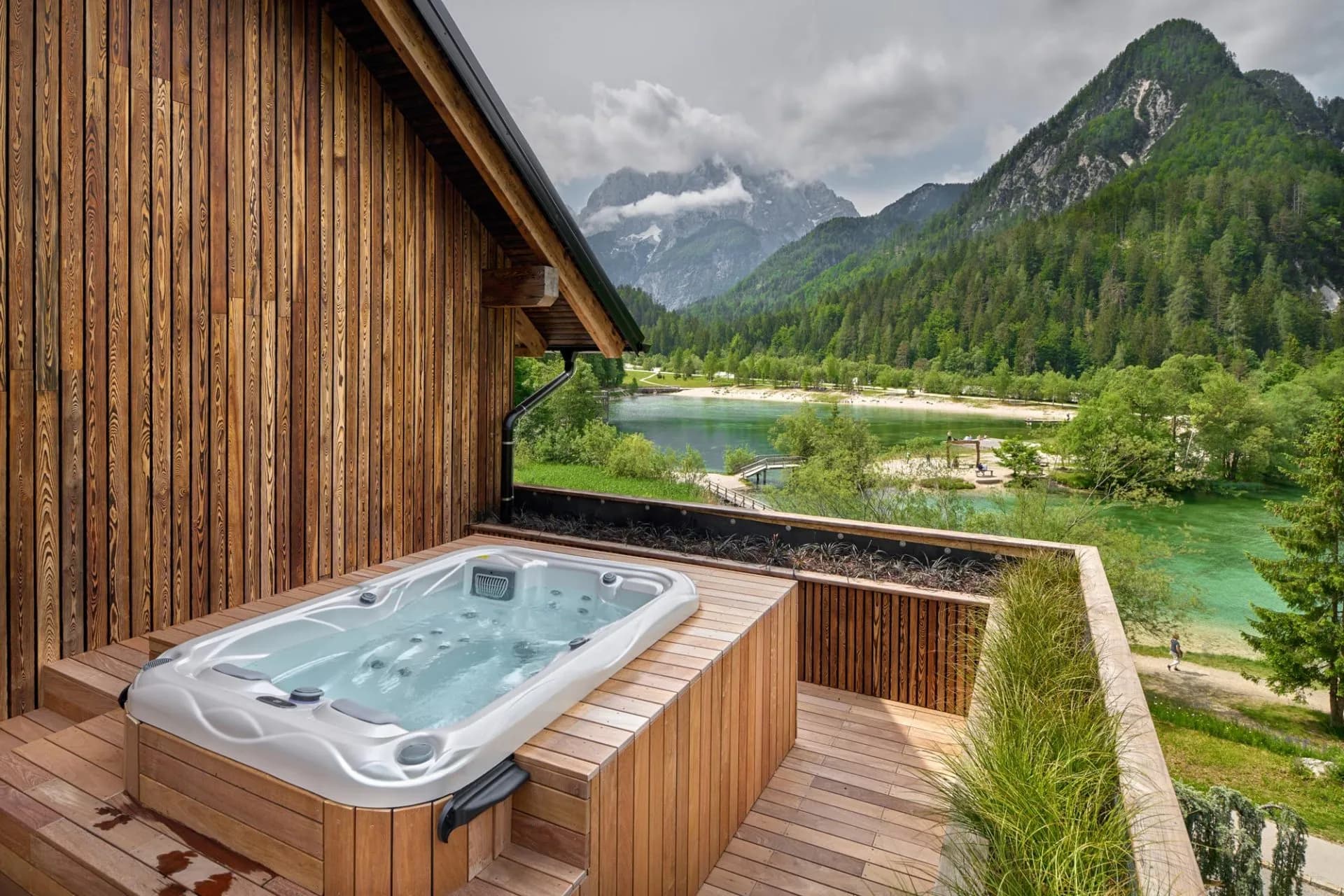 Hot tub on wooden deck overlooking a turquoise lake and forested mountains.