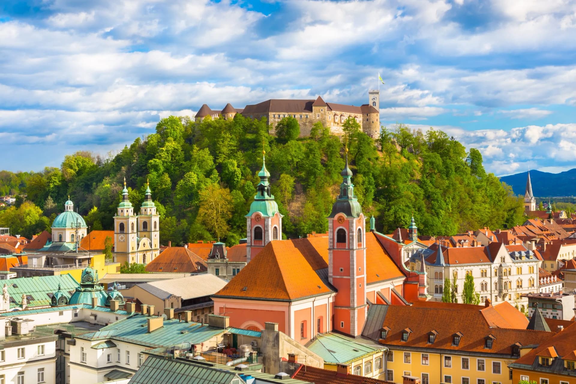 Rooftops of Ljubljana with Franciscan Church and Ljubljana Castle on a green hill.