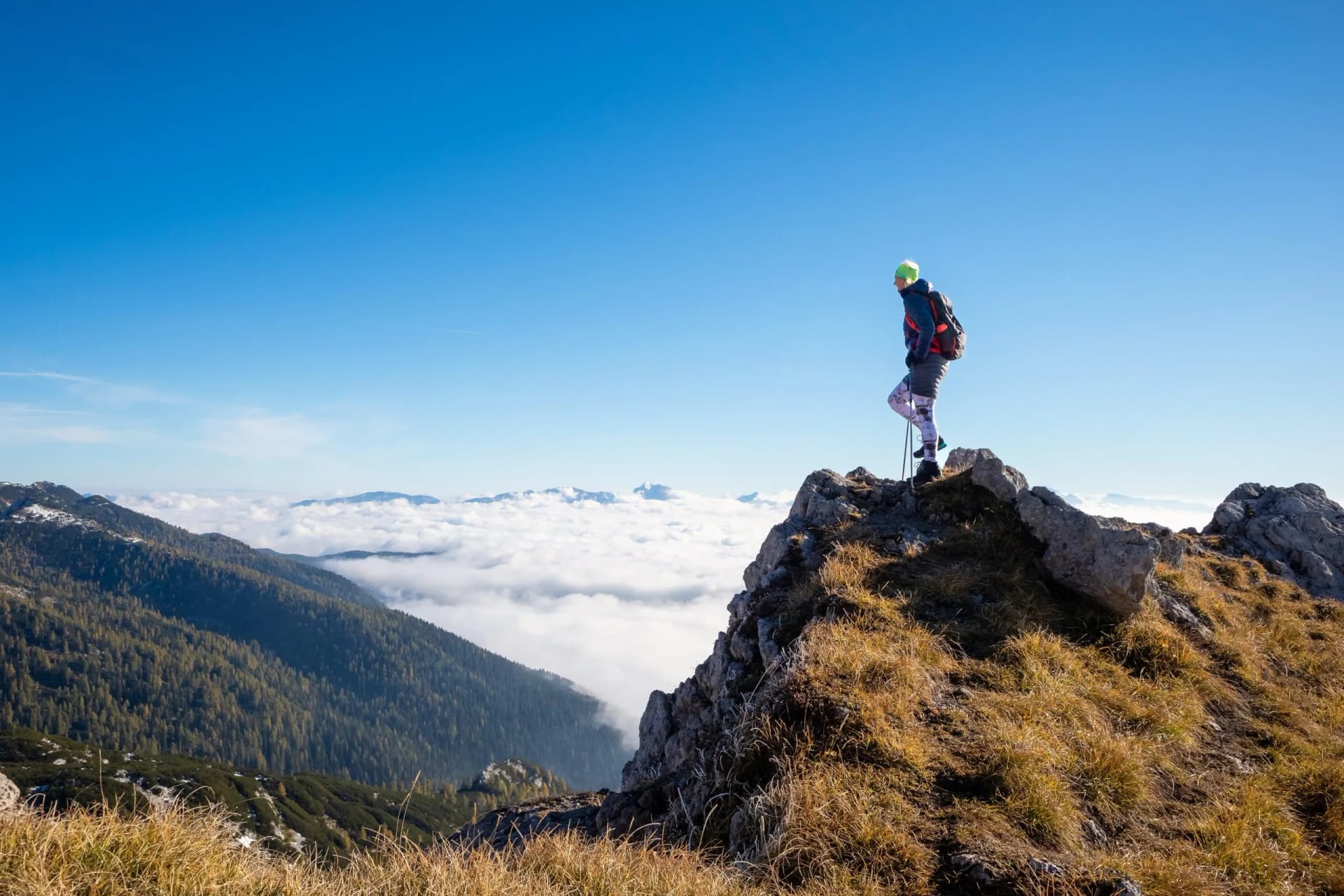 Hiker with poles standing on rocky summit above autumn clouds in Slovenian Alps.
