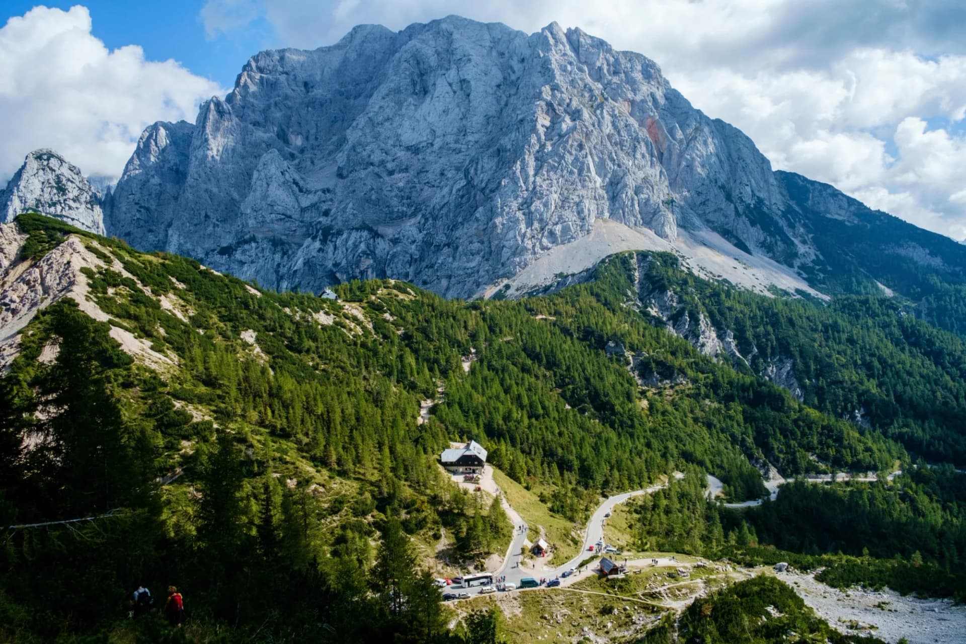 Hikers near a mountain pass with a winding road and alpine lodge beneath a massive gray peak.
