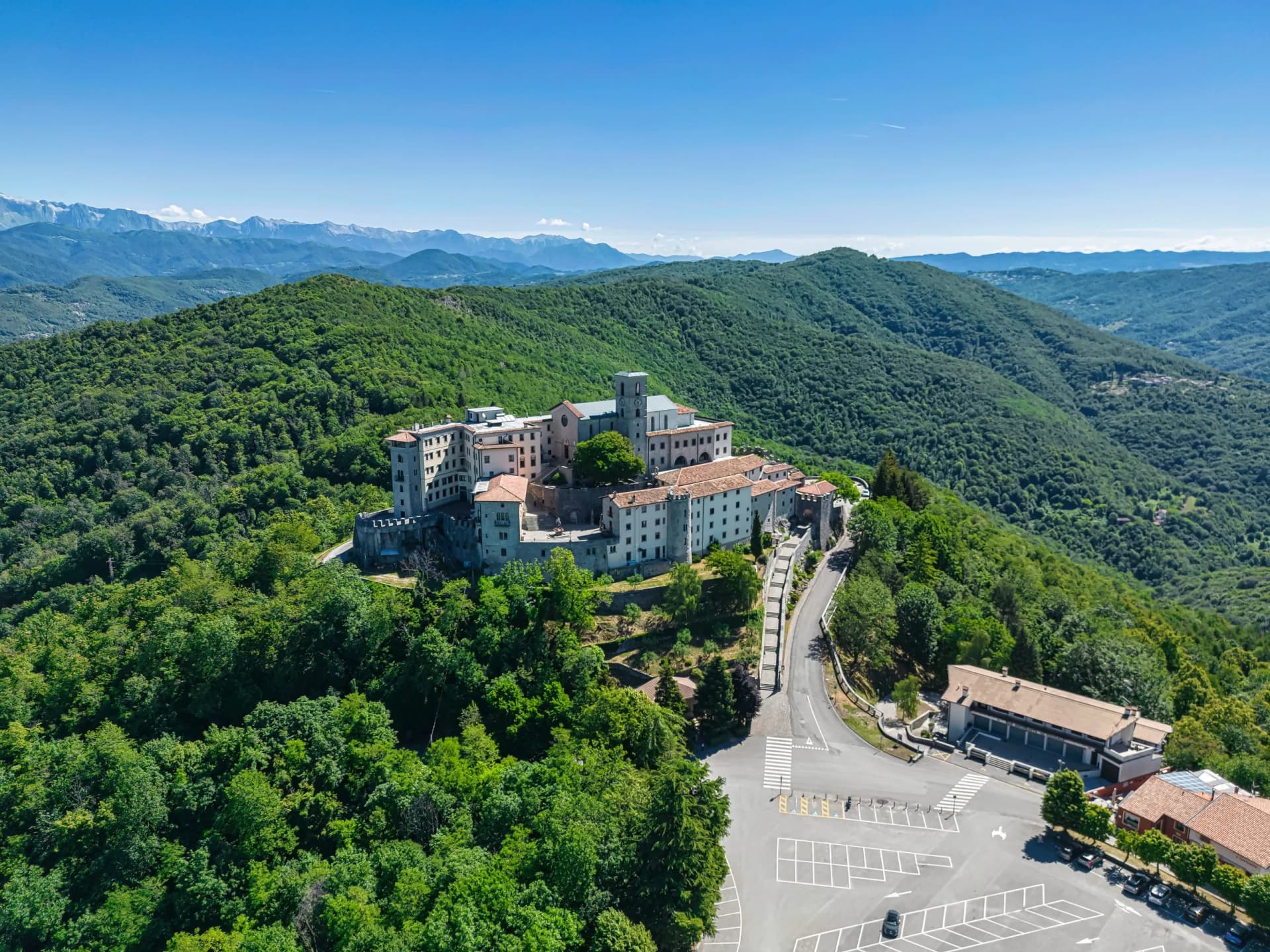 Areal view of the Sanctuary of Castelmonte complex atop a lush green mountain overlooking distant peaks.