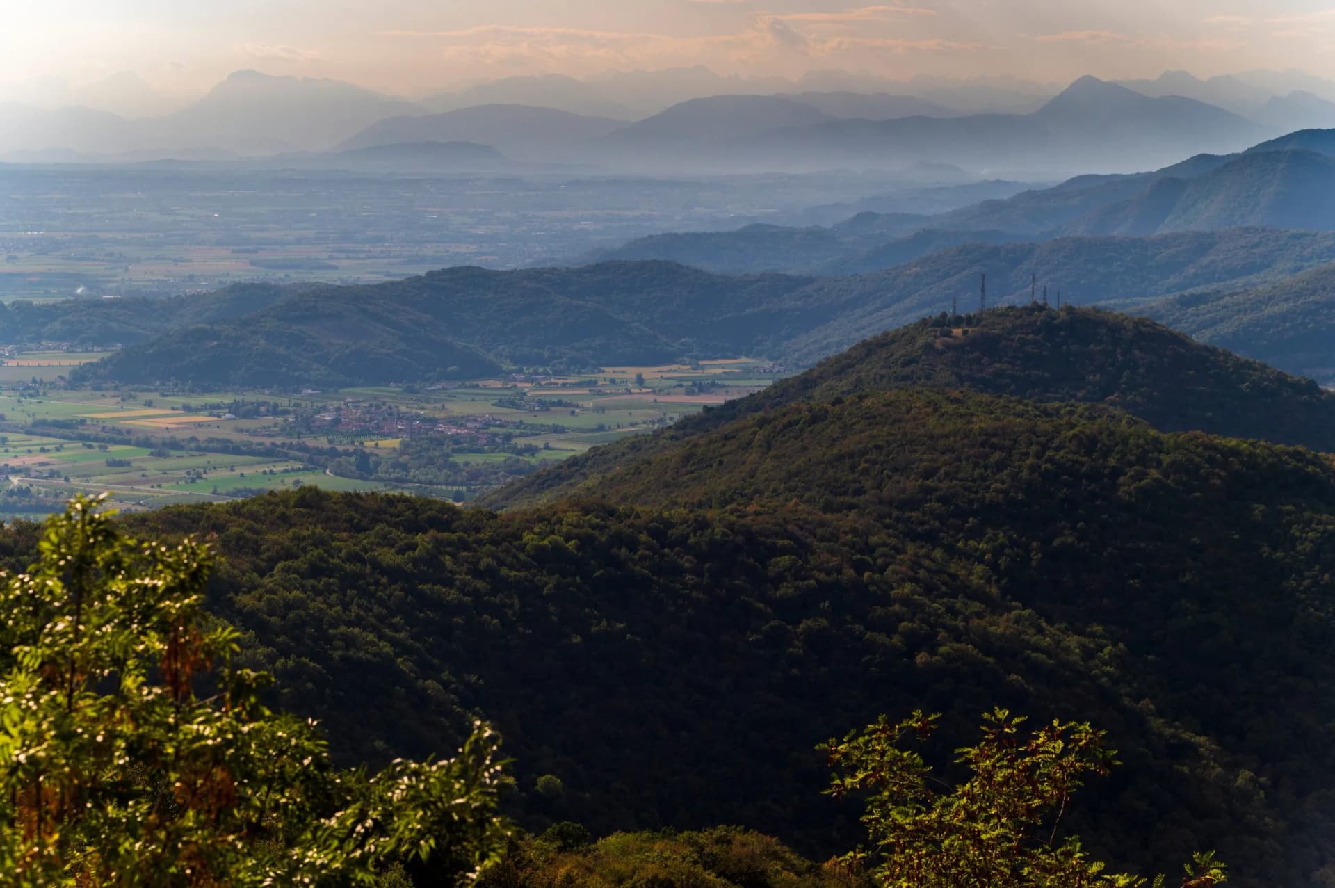 Hazy mountain ranges overlooking a forested hill and valley with a small town, Marian Shrine of Castelmonte.