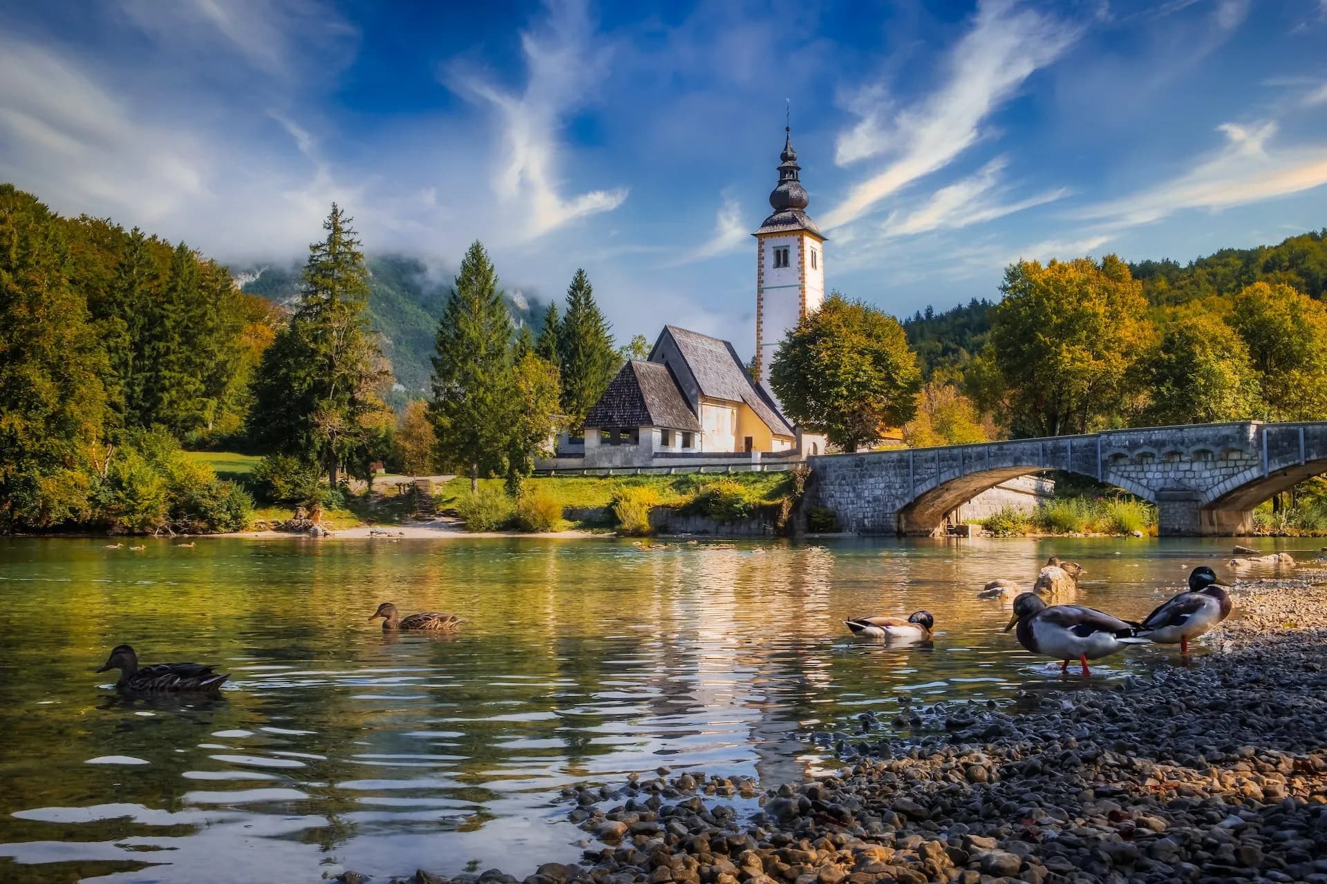 Church with bell tower, stone bridge, and ducks on the water at Lake Bohinj, Slovenia.