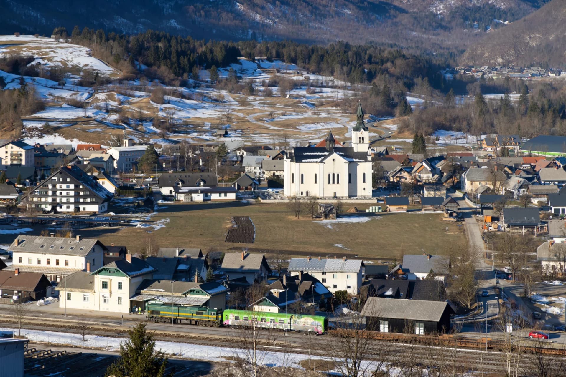 Alpine village of Bohinjska Bistrica with a white church and train in the foreground, winter snow.